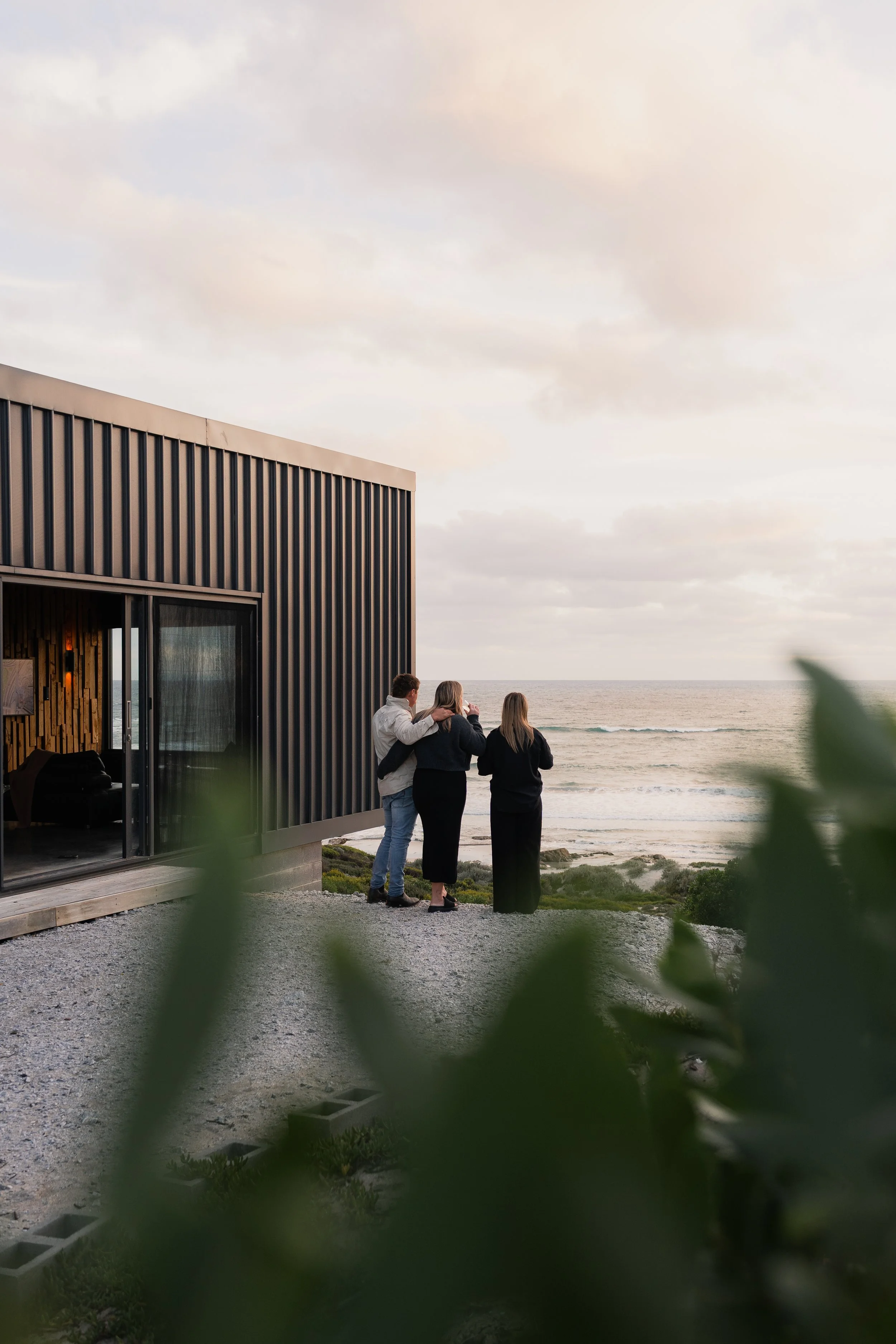 Three people standing together near the beach, gazing at the ocean during sunset, with a modern building on the left side of the image.