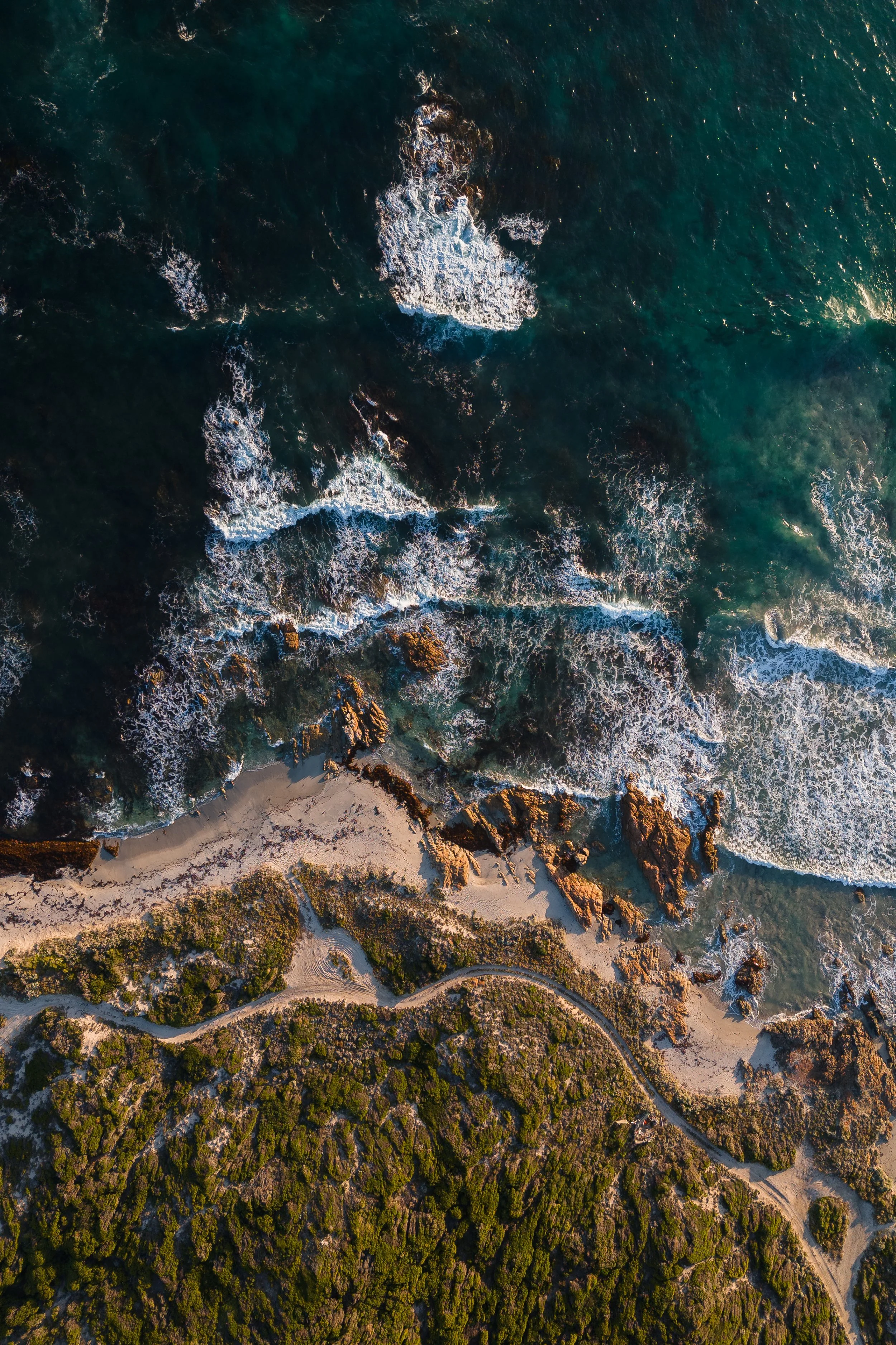 Aerial view of a coastline featuring rocky cliffs, green vegetation, sandy beach, and the ocean with waves.