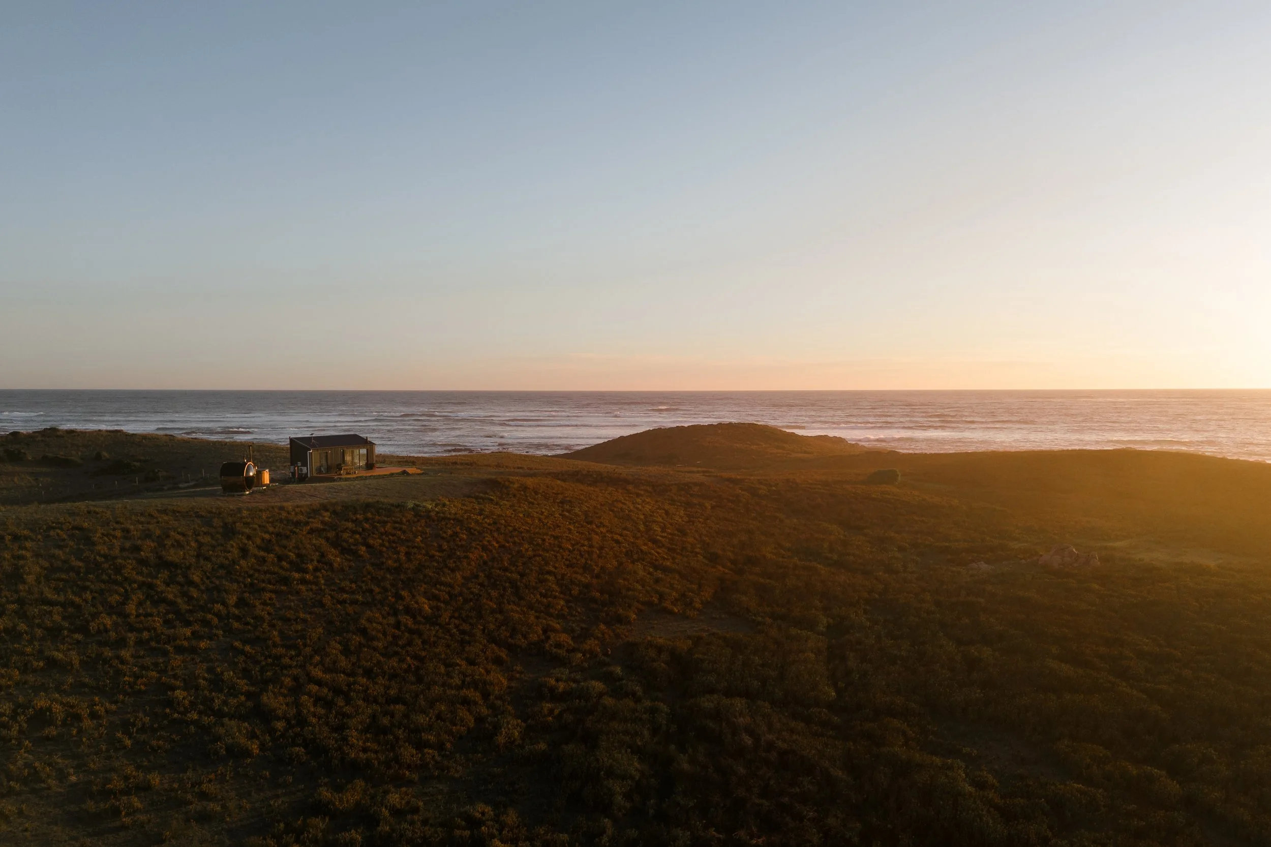 Sunset over a beach with low waves, grassy dunes in the foreground, and structures including a small building and tanks on the dunes.