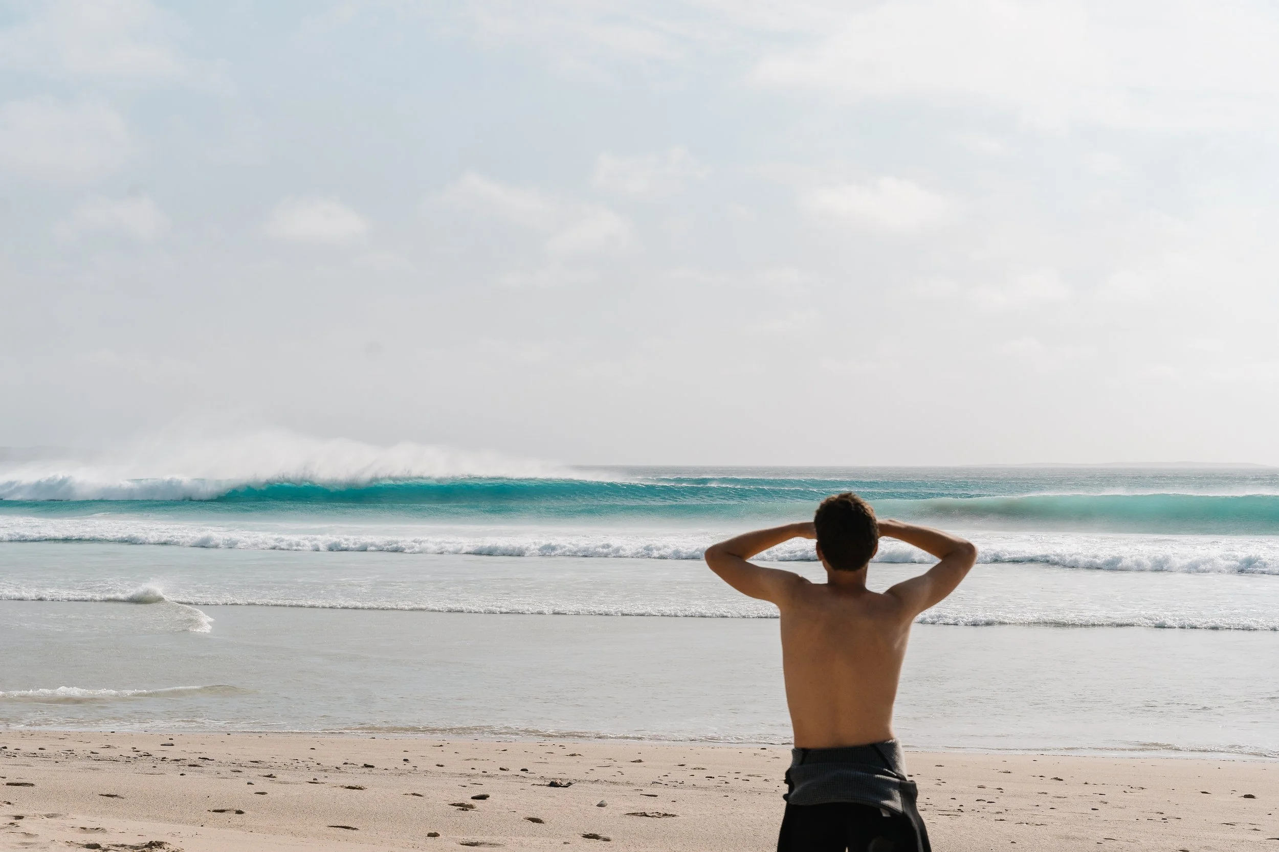 A shirtless man standing on a sandy beach, facing the ocean with his hands resting on his head. The waves are crashing in the distance under a cloudy sky.