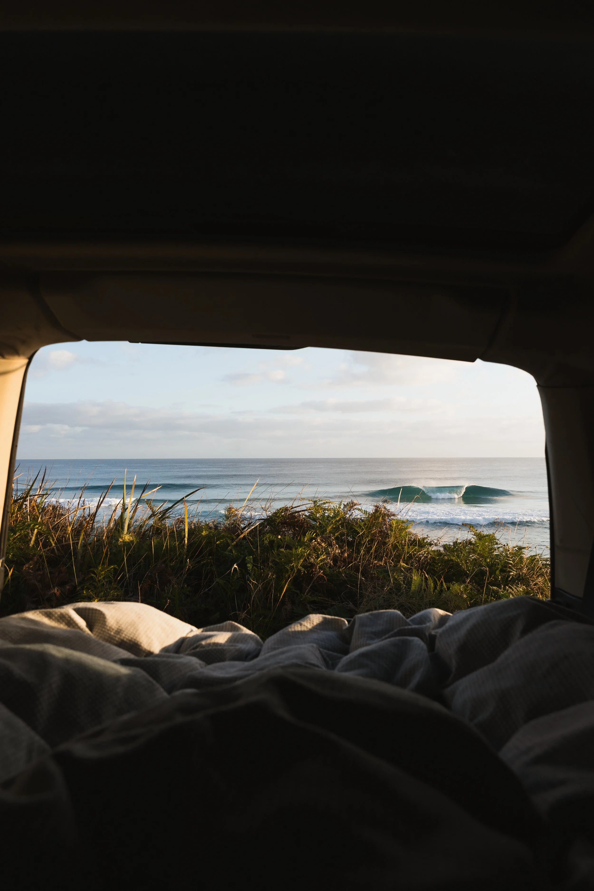 View of the ocean and waves from inside a vehicle, with part of the bed or sitting area visible in the foreground, and the sky with clouds in the background.