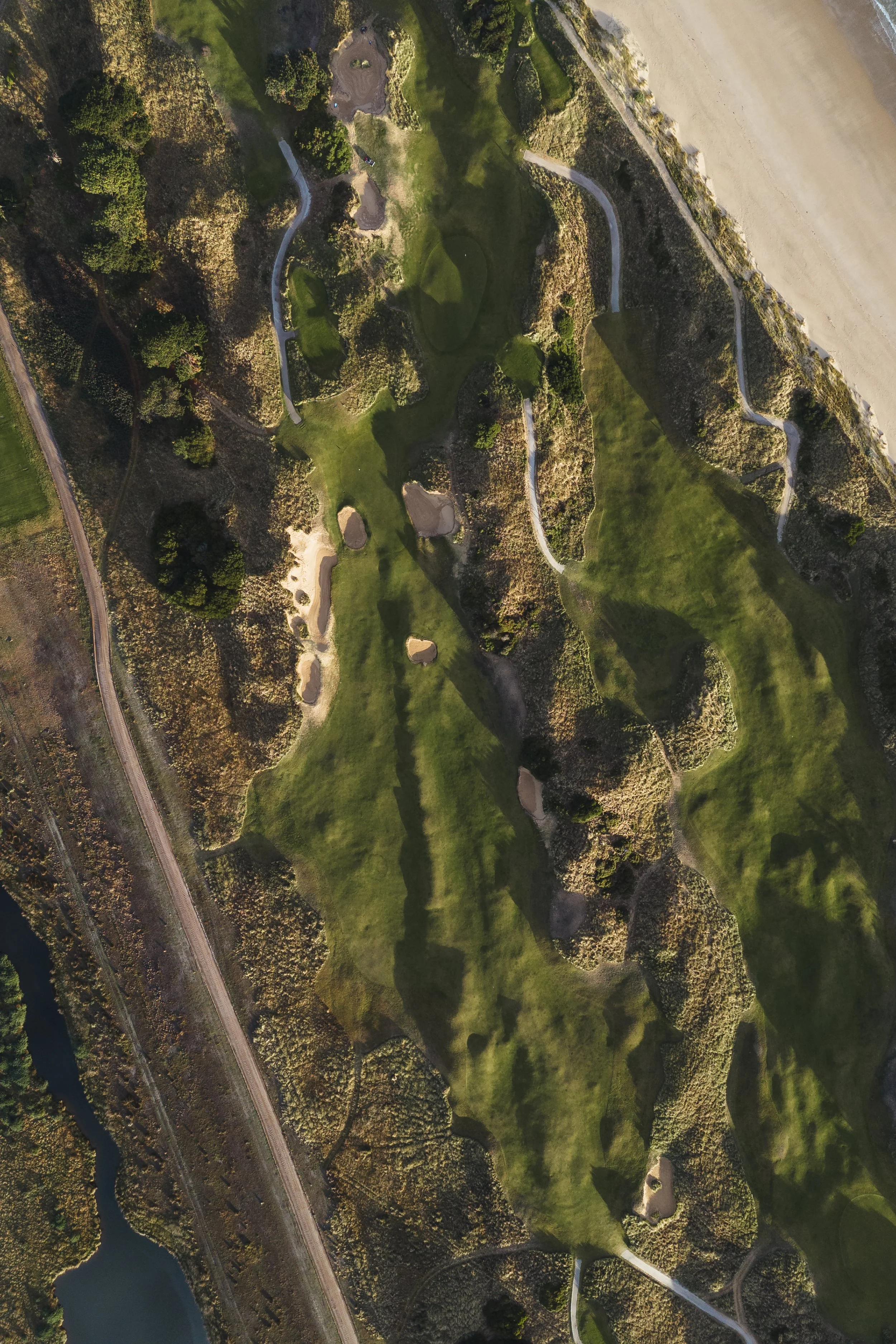 An aerial view of a golf course with winding pathways, sand traps, and lush greenery, bordered by trees and a beach on one side.