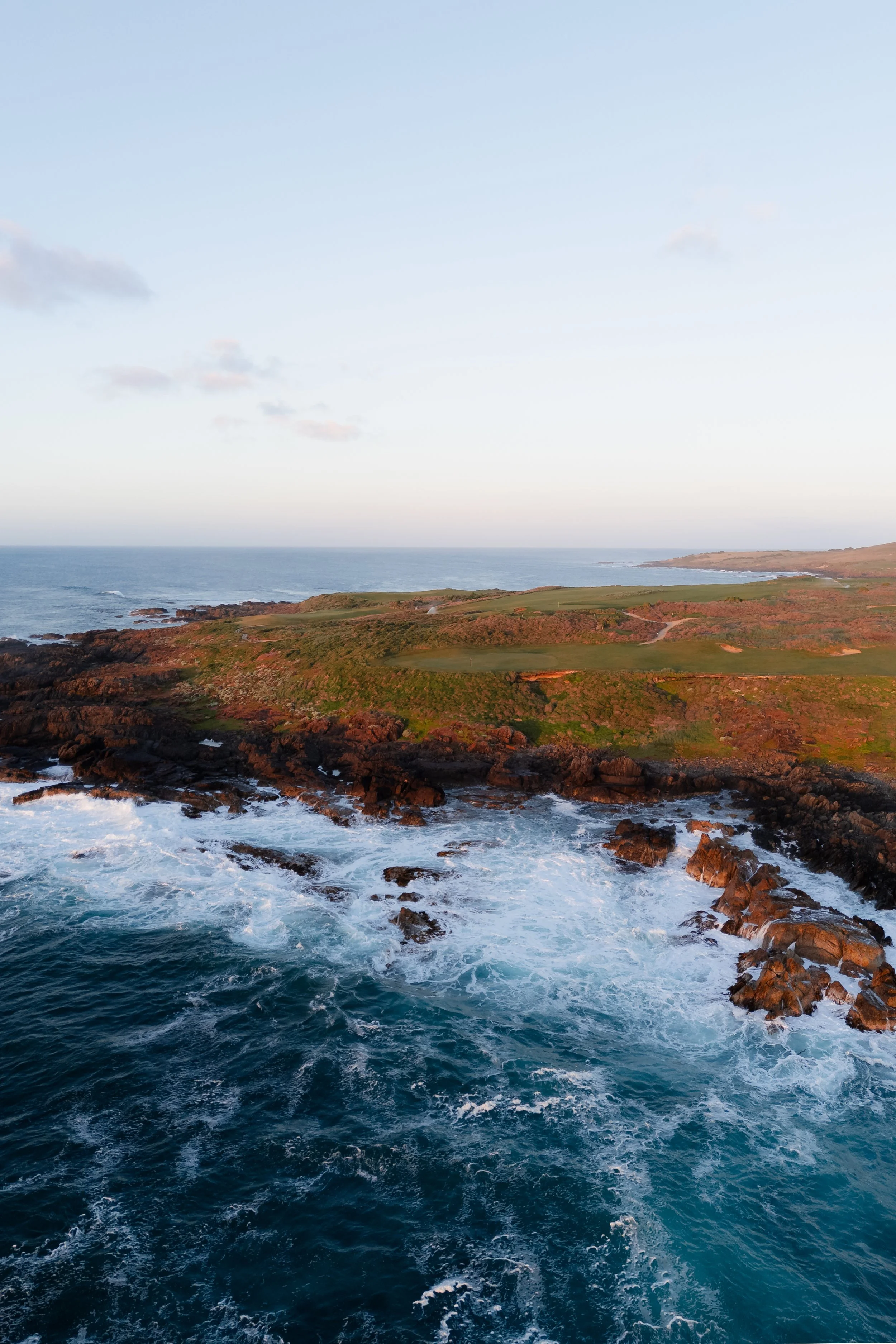 Coastal view with rocky shoreline, green grassy areas, and the ocean waves crashing against rocks under a mostly clear sky.