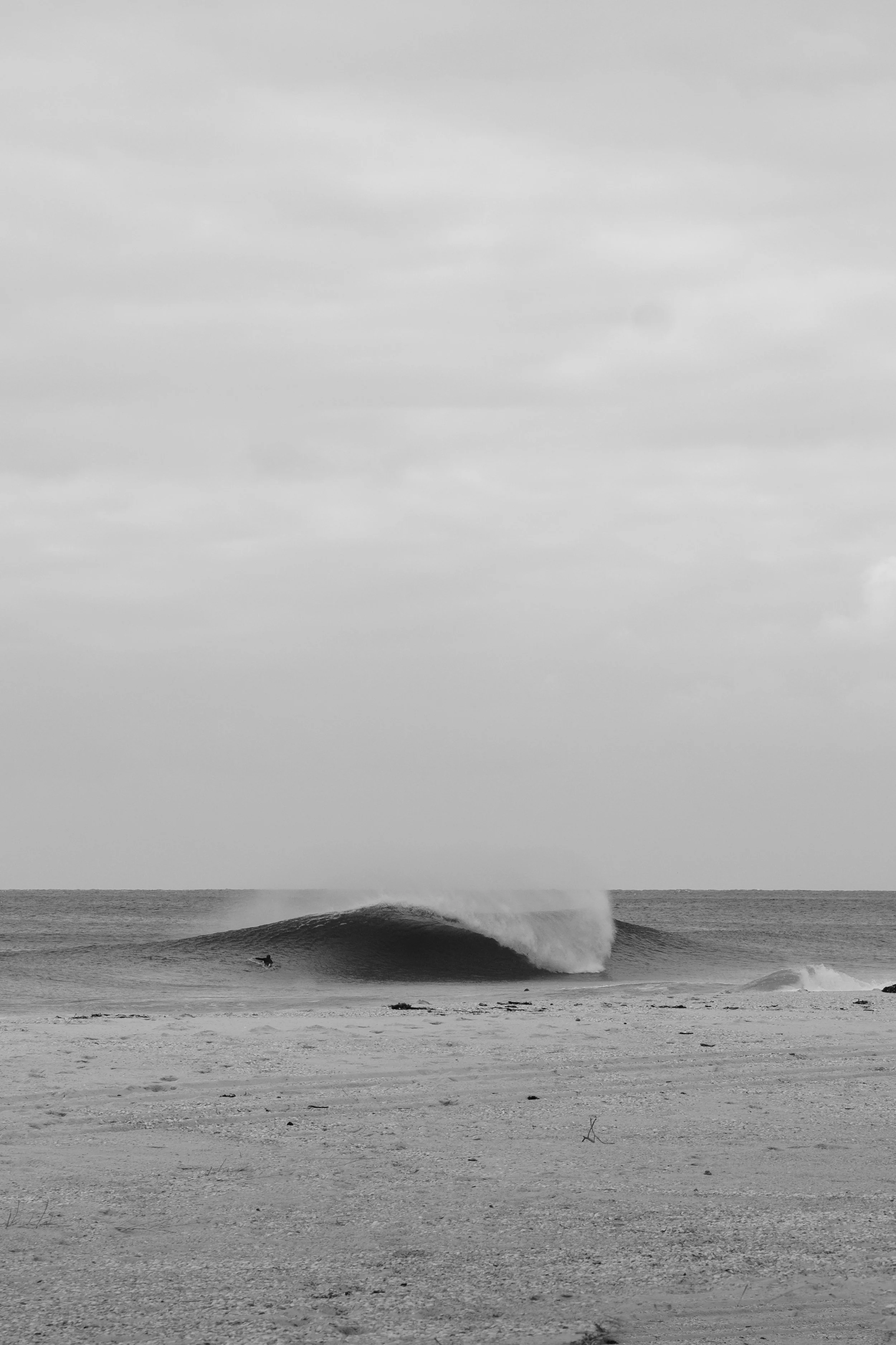 Black and white photograph of a beach with a large, powerful wave breaking, and a surfer riding it in the distance.