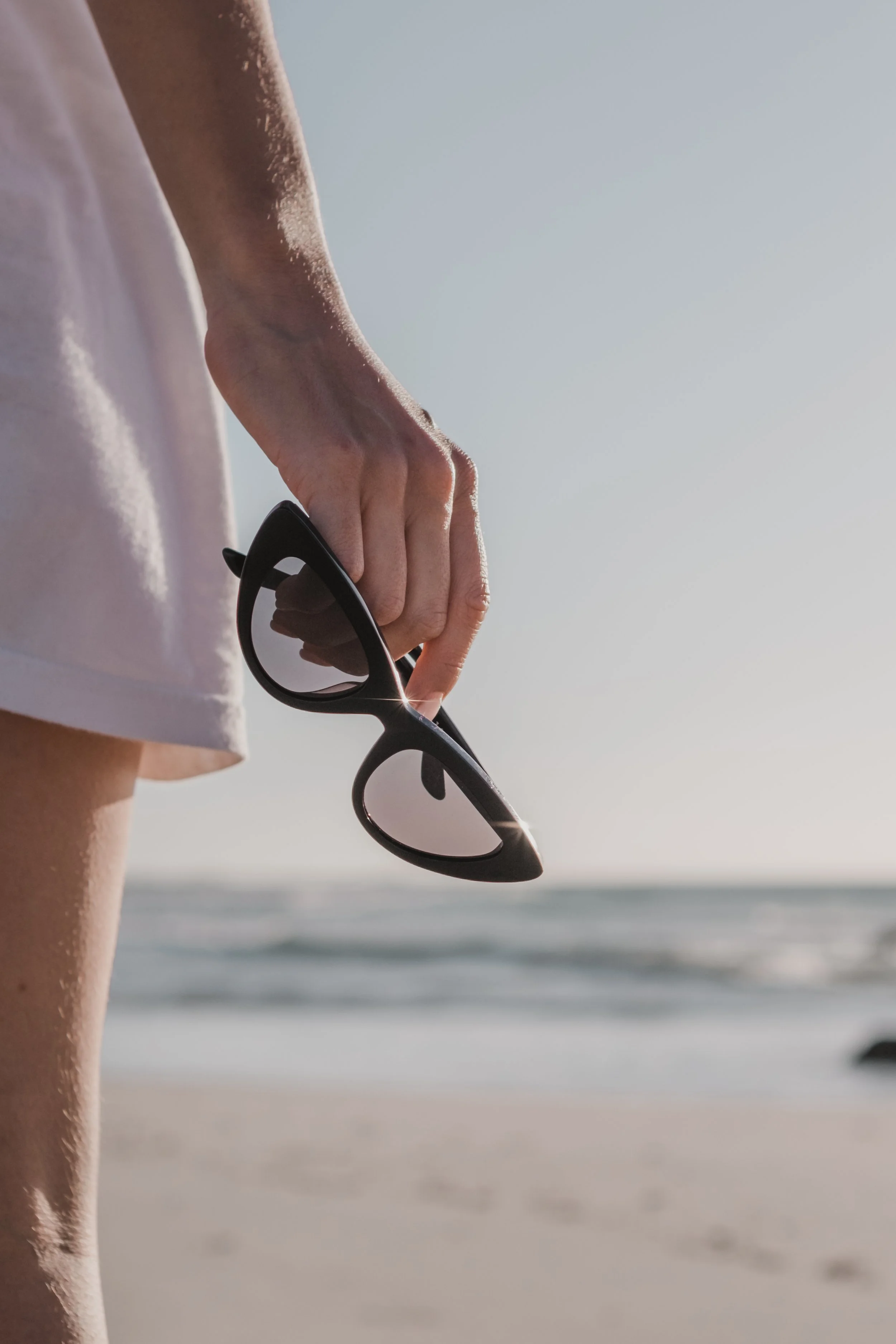 Close-up of a person’s hand holding sunglasses with a beach and ocean in the background.
