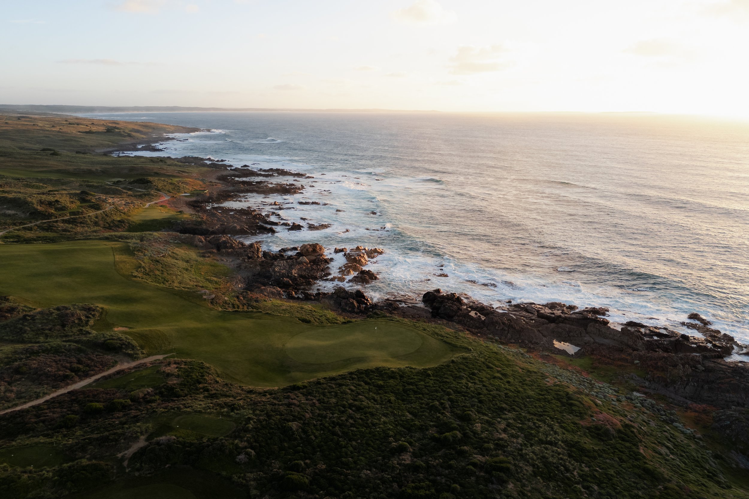 Aerial view of a coastline with green golf course grounds and rocky shores, extending into the ocean under a clear sky with the sun setting on the horizon.