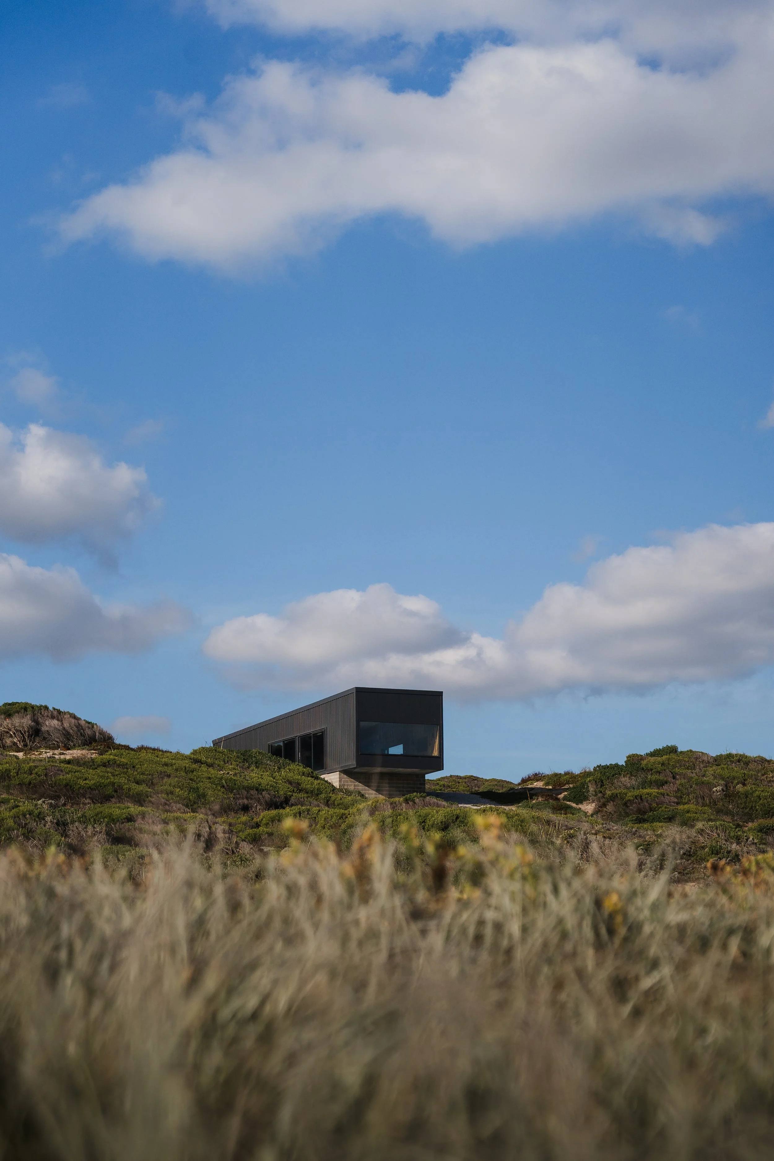 Modern black house on grassy hill with a blue sky and scattered white clouds.