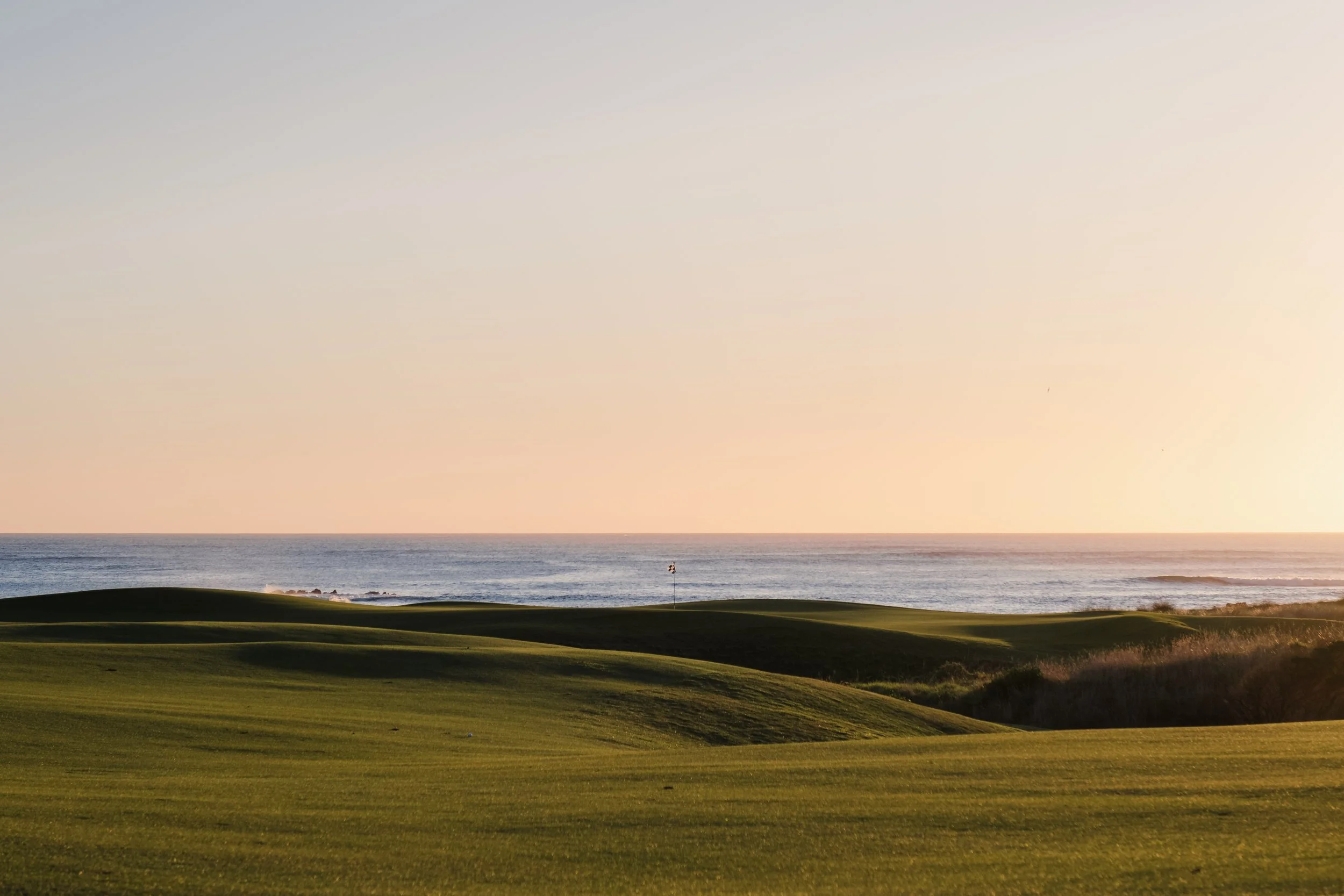 A view of a golf course with the ocean in the background, captured during sunset with the sky displaying soft pastel colors.