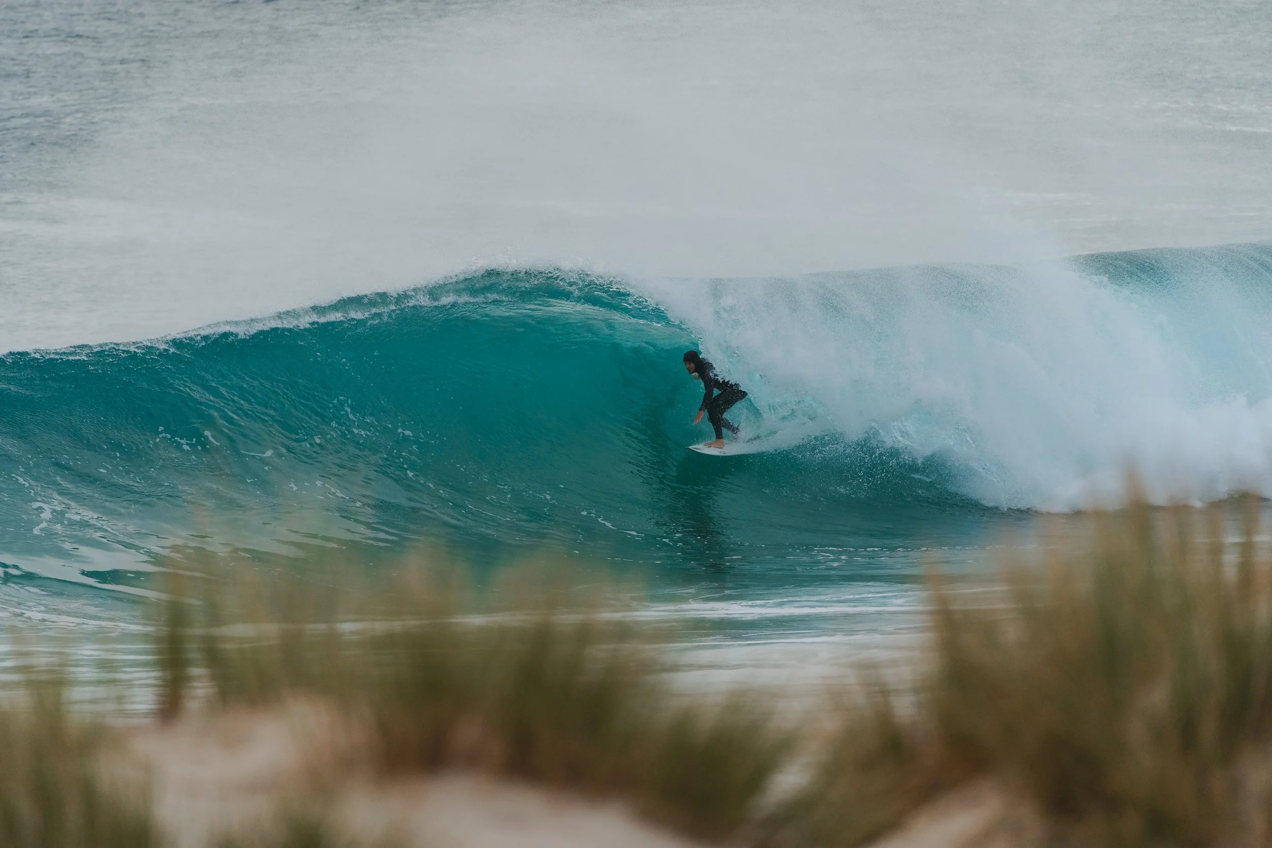 A person surfing on a large turquoise wave near a beach, with the image taken from a low angle through the sand.