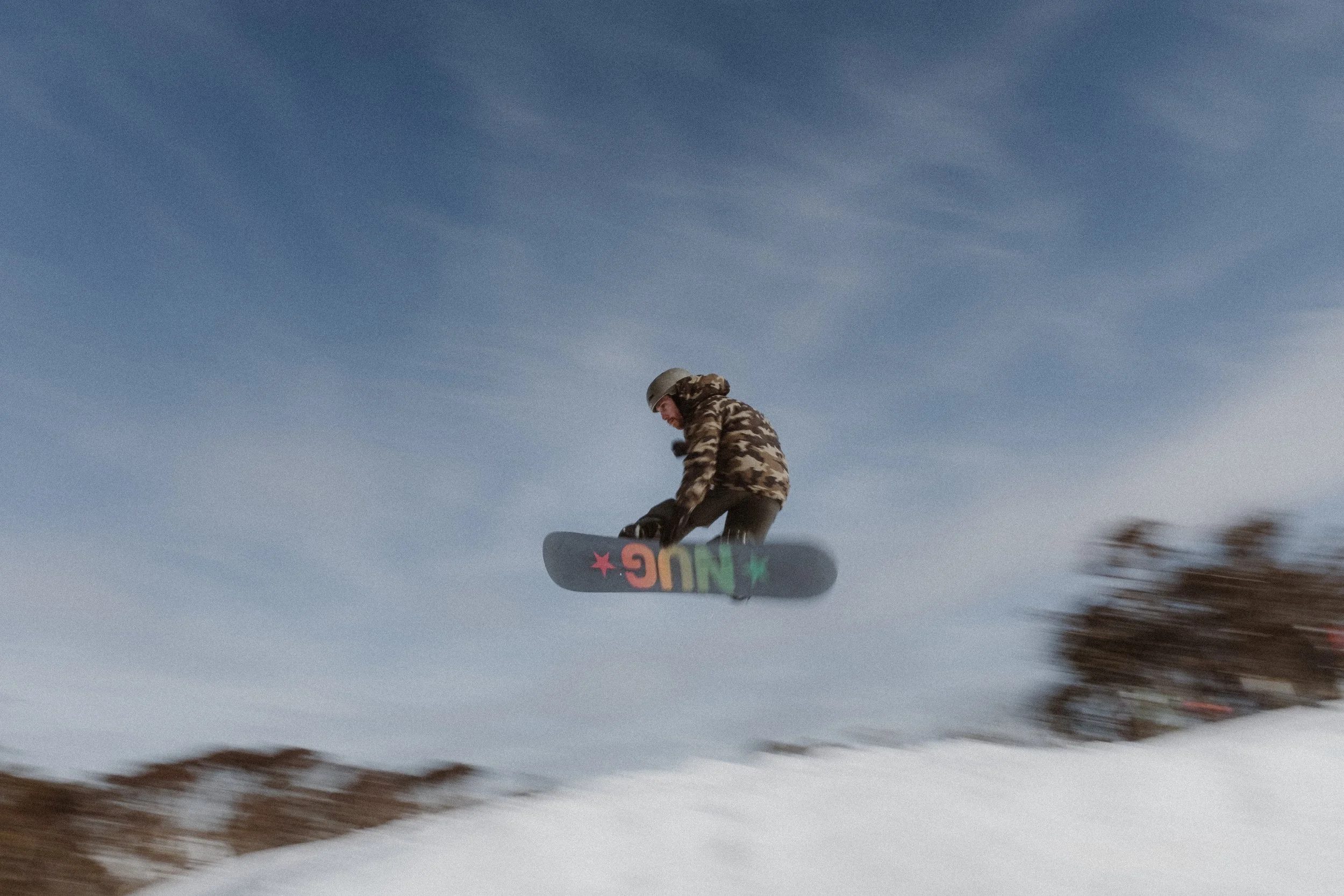 A person wearing camouflage clothing and a helmet snowboarding on a snowy slope against a clear blue sky.