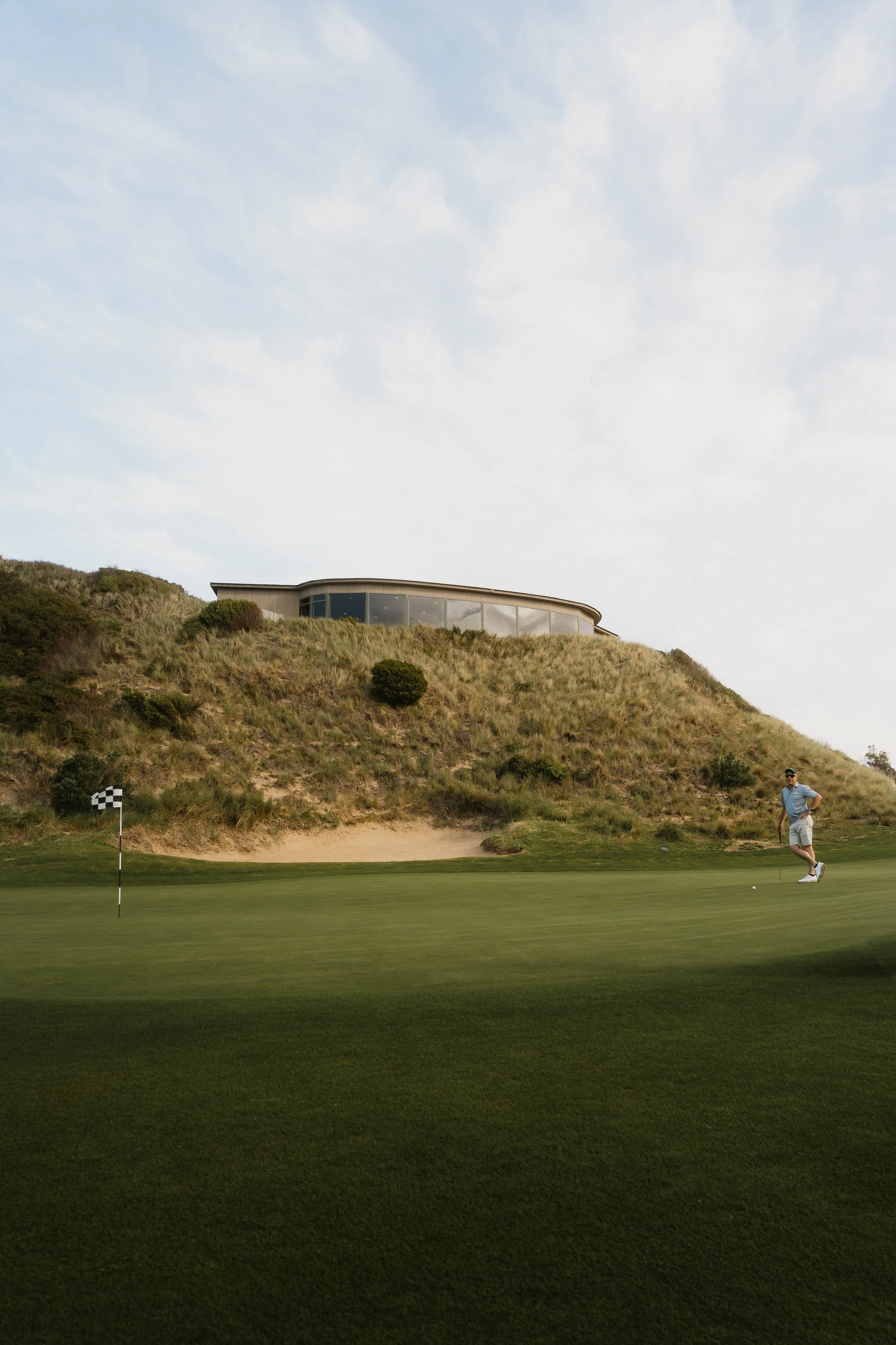 A golfer on a golf course preparing to putt near the hole with a flagstick. There is a hillside with grass and bushes behind him, and a modern building with glass windows atop the hill. The sky is partly cloudy.