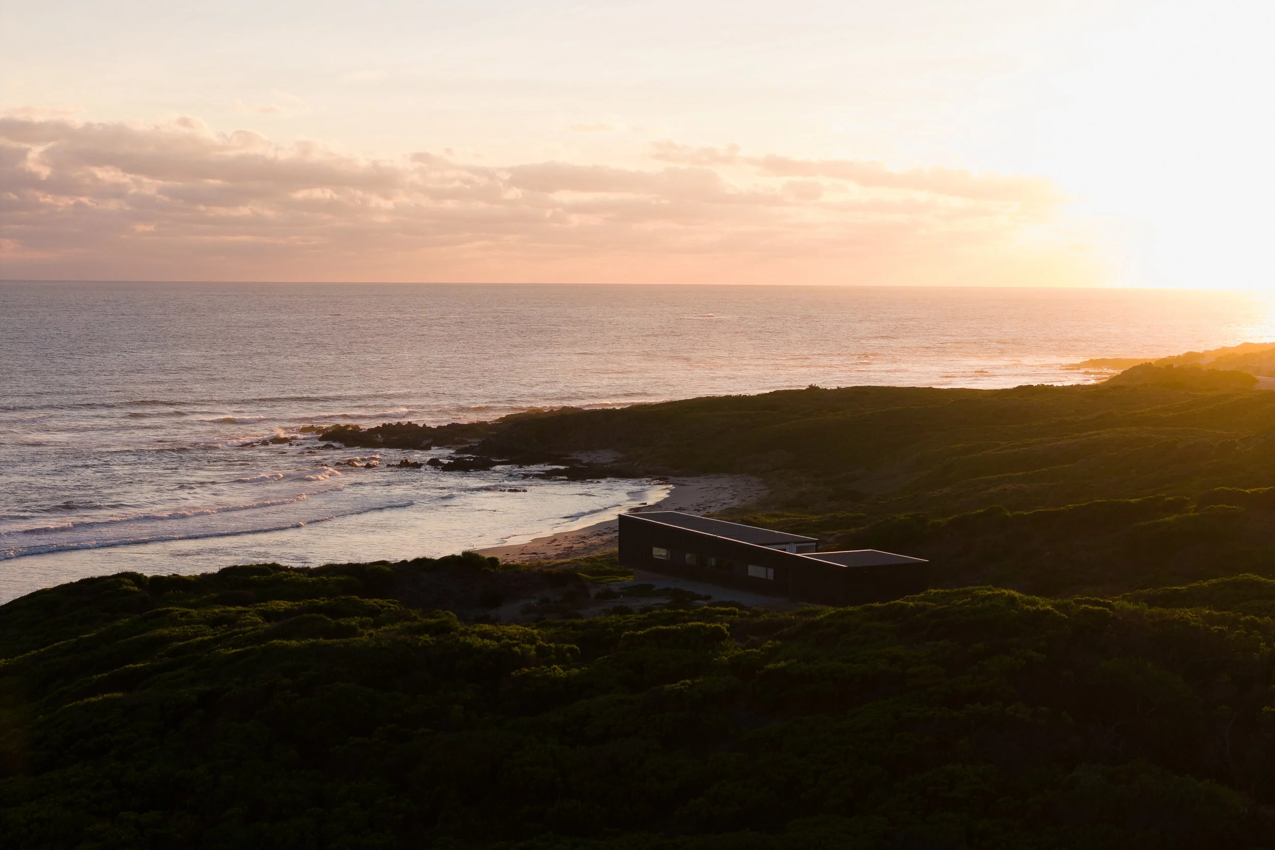 Sunset over the ocean with a rocky shoreline and a modern house in the foreground, surrounded by green vegetation.