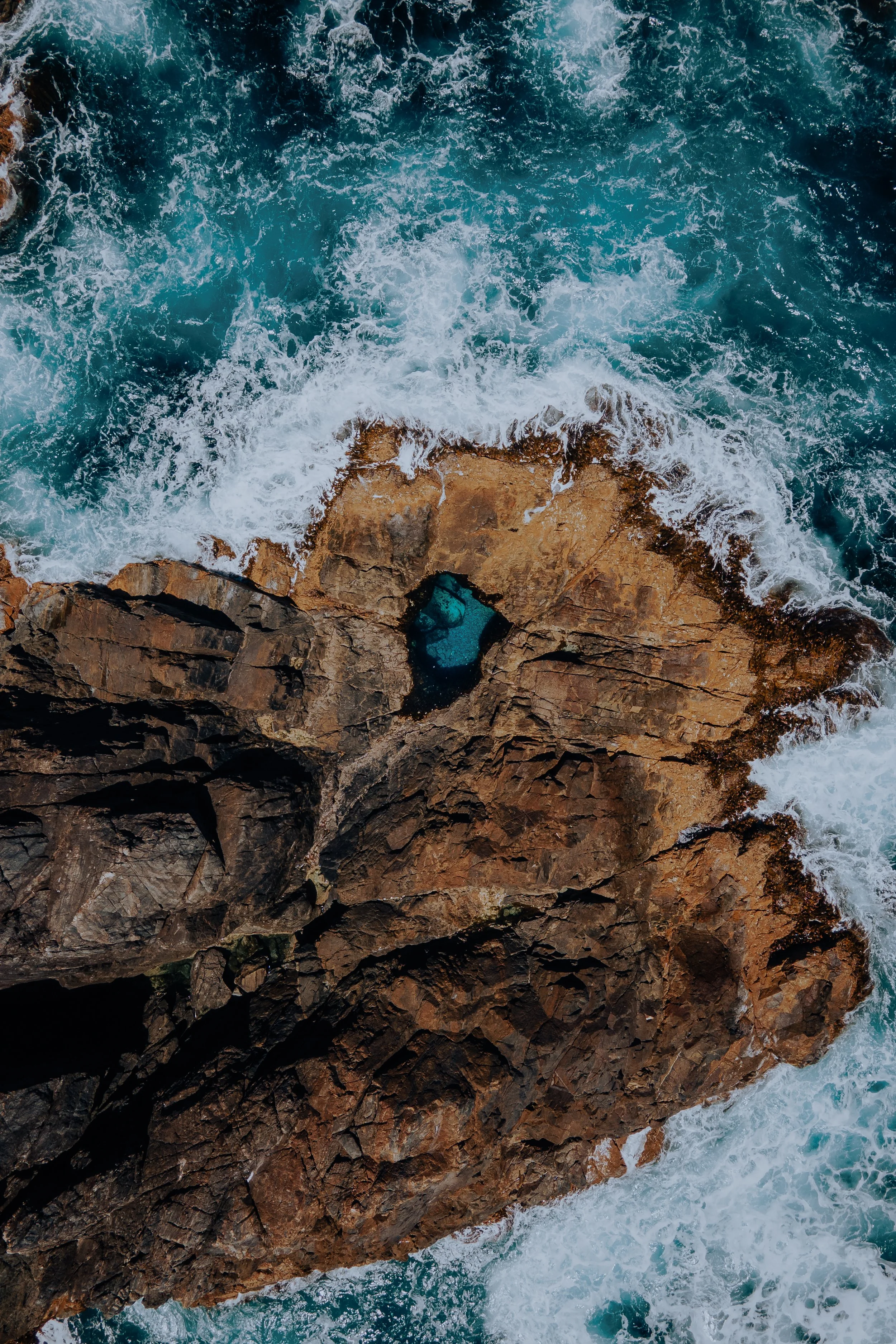 Overhead view of ocean waves crashing against a rocky shoreline with a small pool of water.