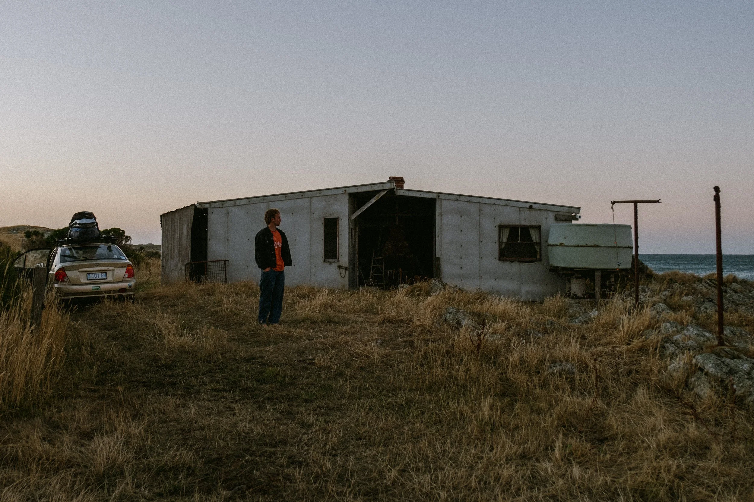 Person standing outside near an old, weathered trailer by the sea during sunset or dusk, with a parked car and a backpack on its roof.
