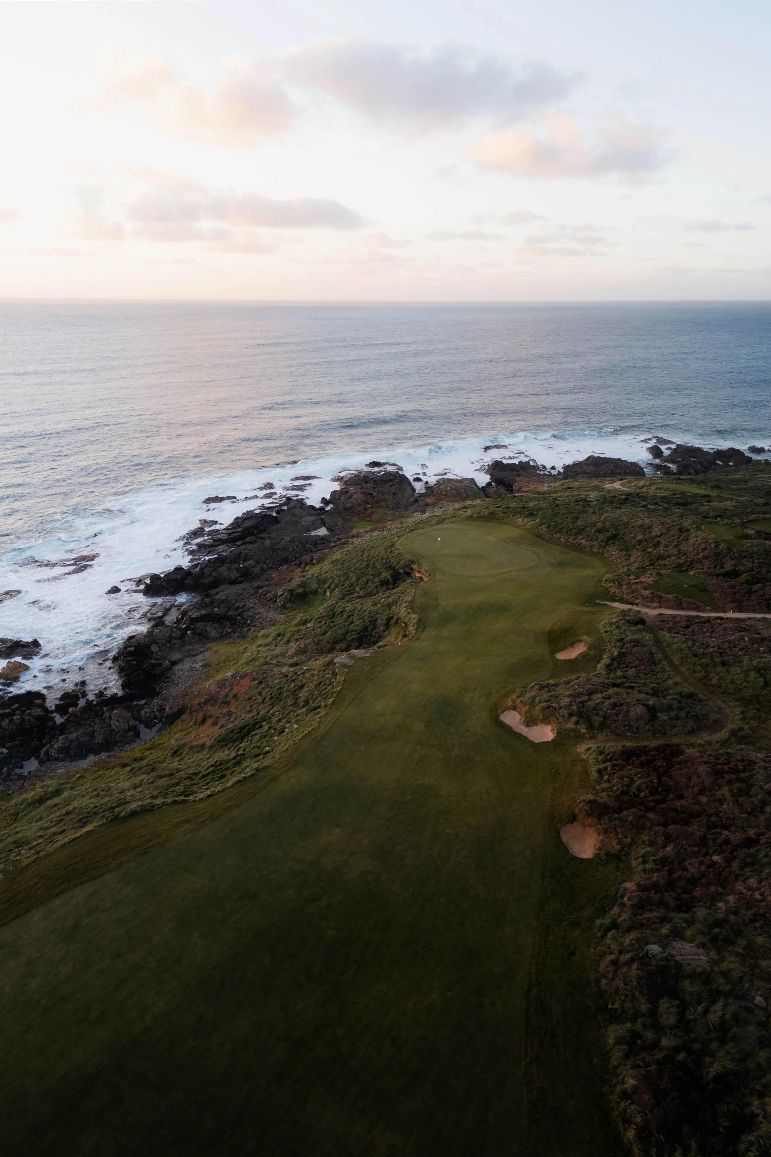 A scenic view of the ocean with waves crashing against rocky shoreline and a green golf course with sand bunkers in the foreground, under a sky with scattered clouds.