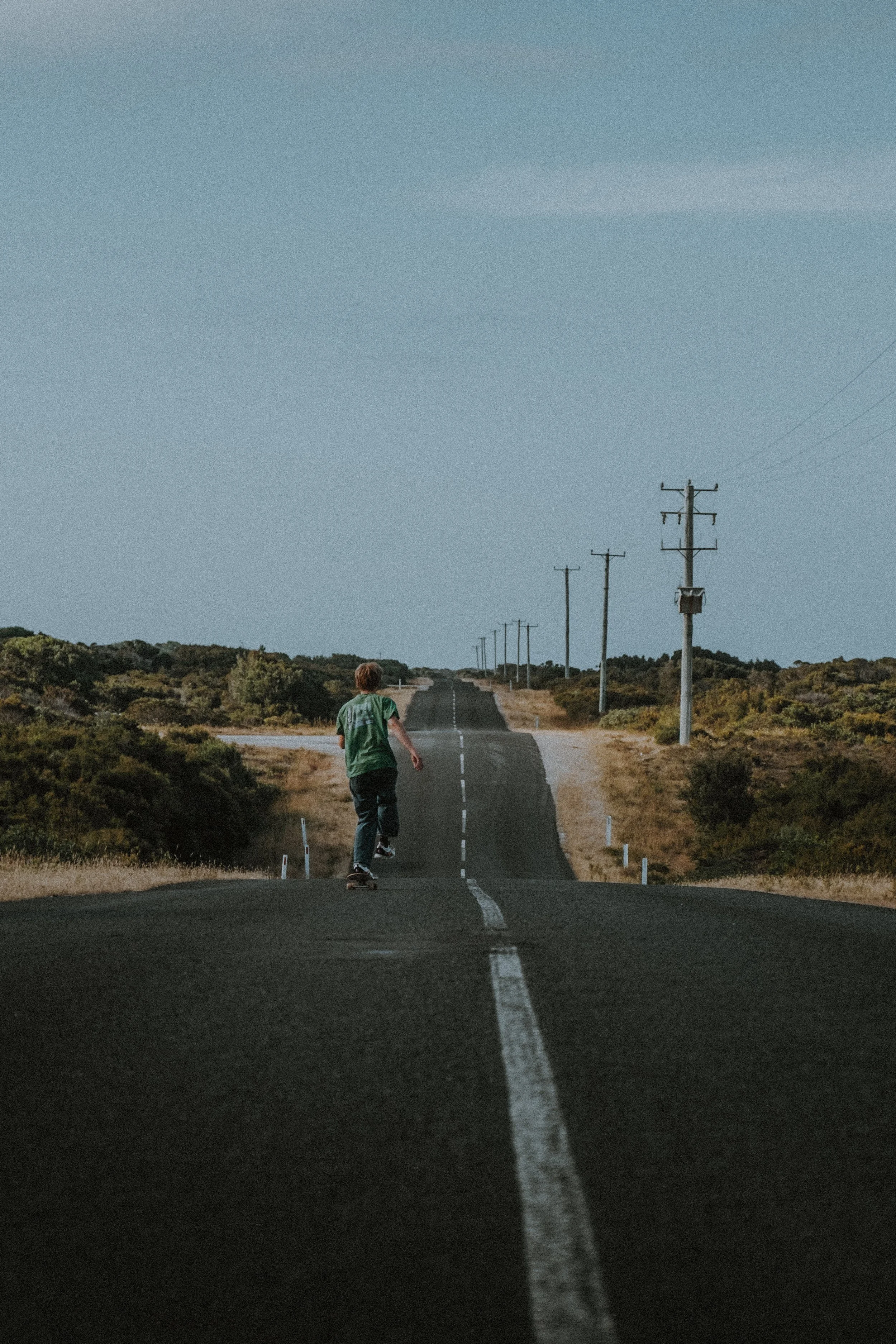 Young person skateboarding on a deserted rural road with telephone poles and overgrown vegetation, under a clear blue sky.