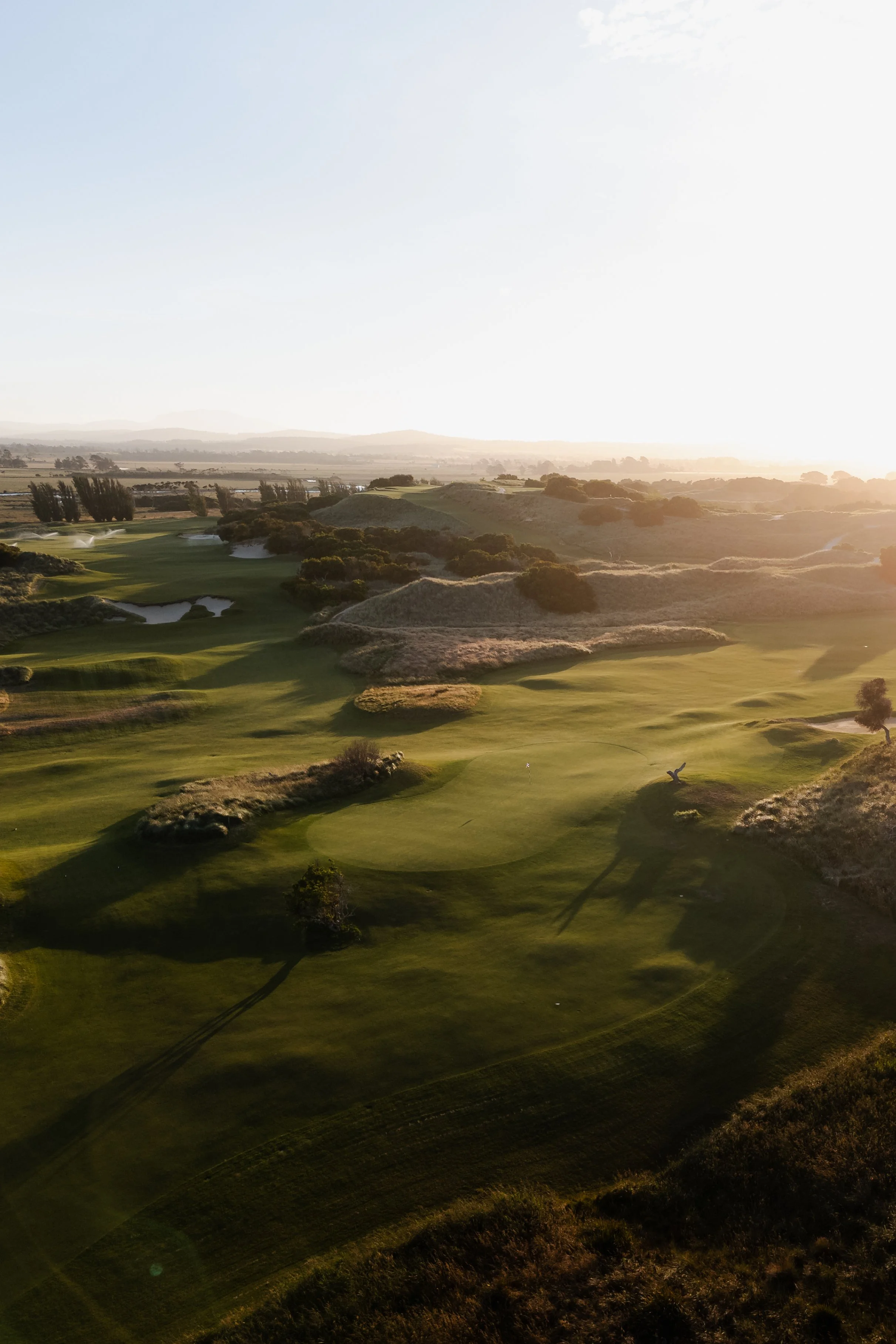 A golf course during sunset with lush green fairways, sand traps, and scattered trees, under a clear sky with the sun low on the horizon.