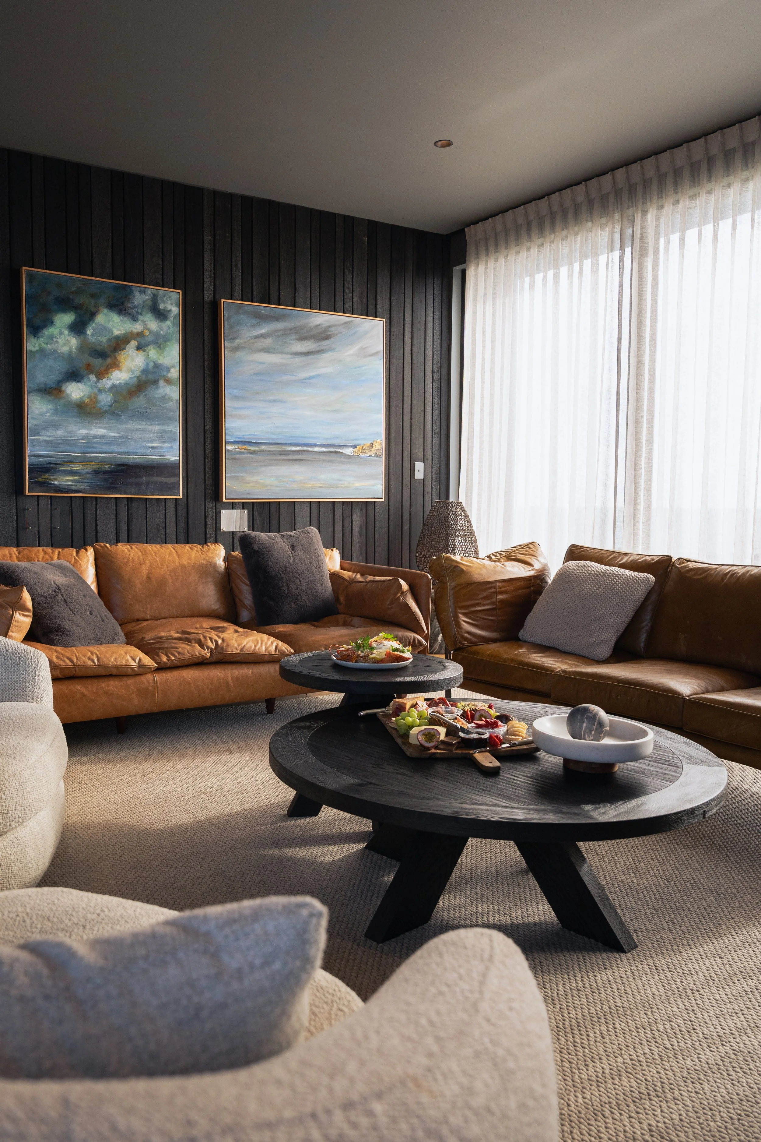 A living room with beige sofa and coffee table, dark wood wall with two landscape paintings, and white sheer curtains allowing light to enter.