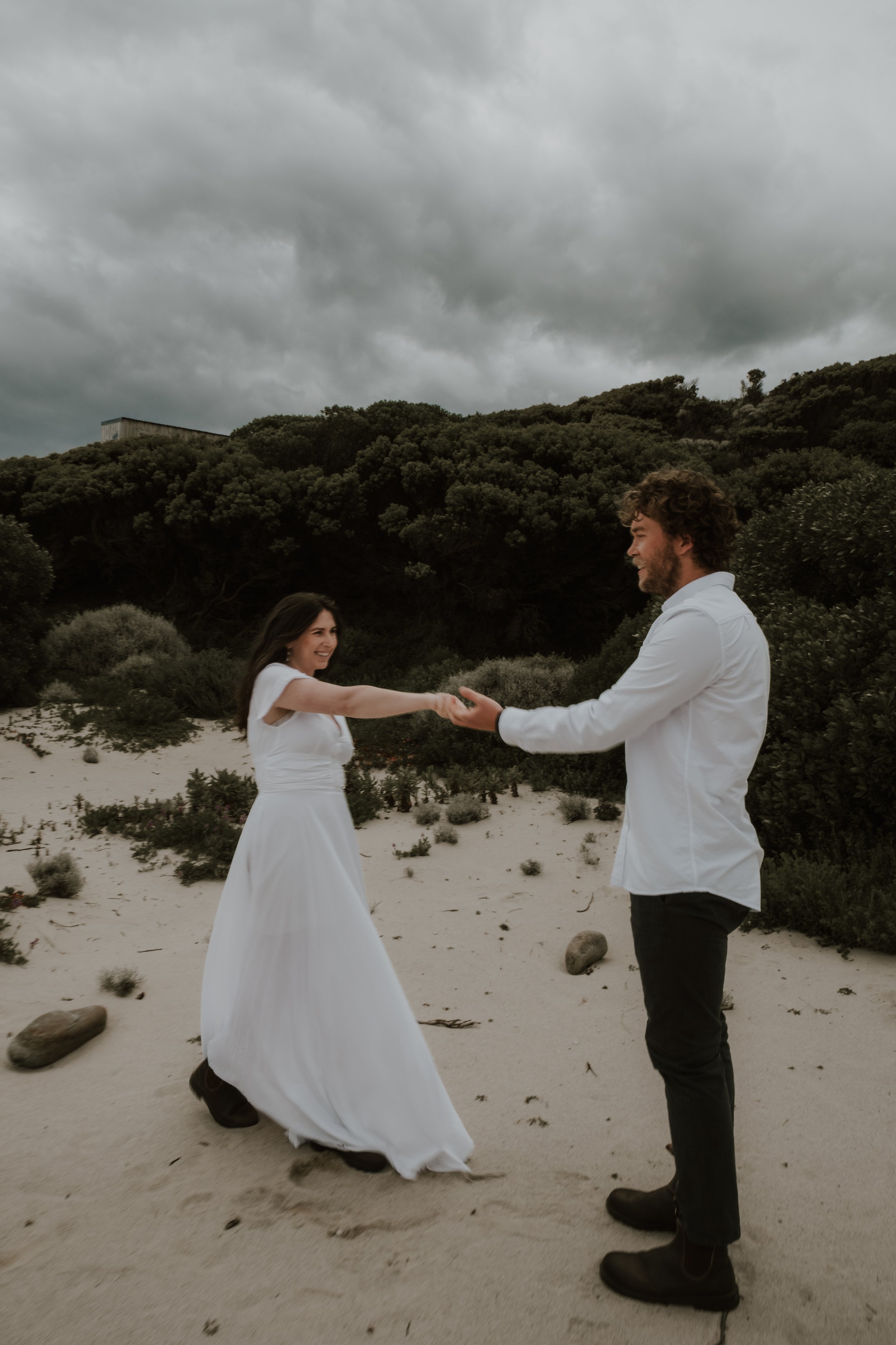 A couple dressed in white, holding hands and smiling at each other on a sandy beach with cloudy sky and green hills in the background.
