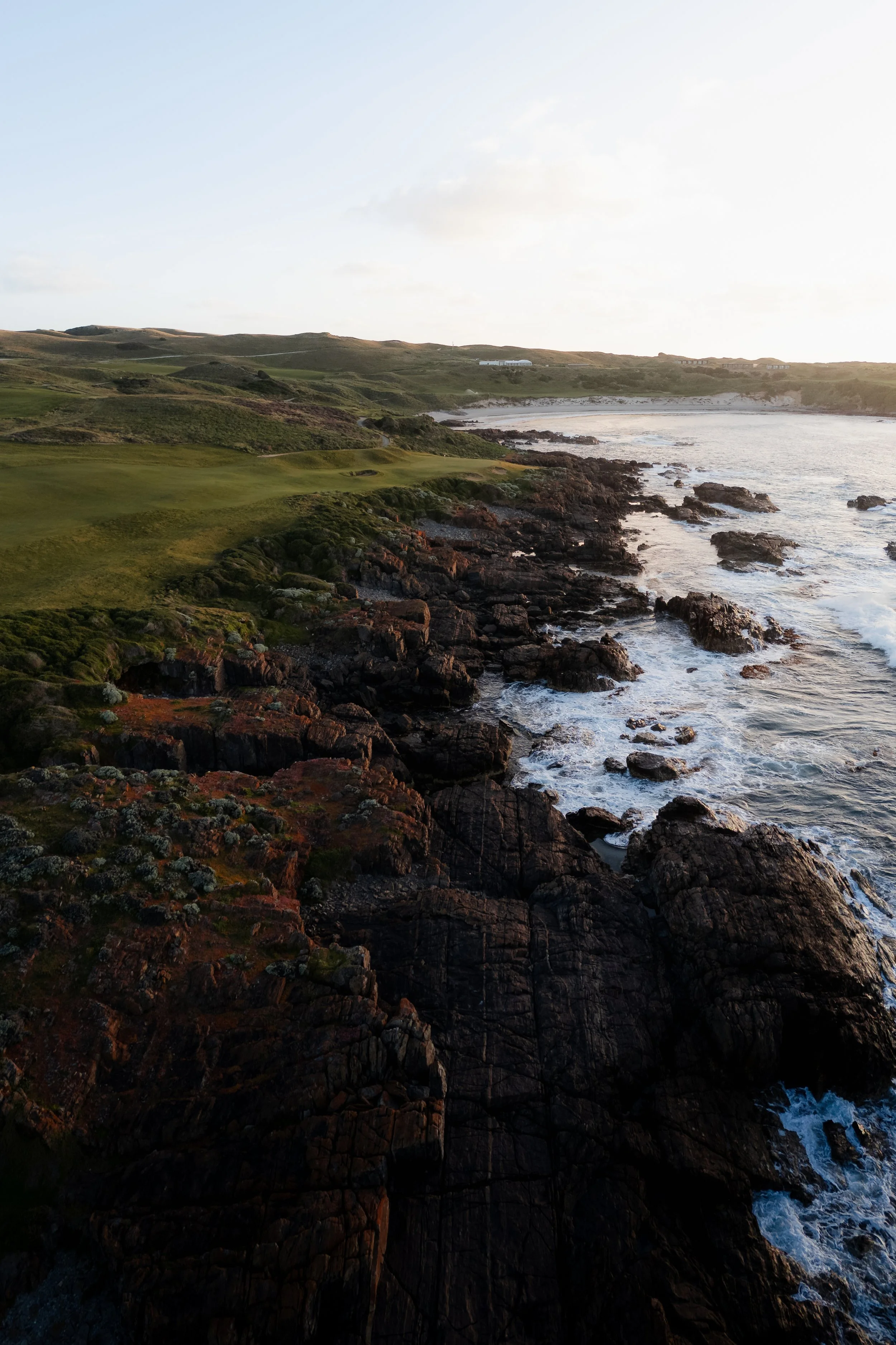 Coastal landscape with green hills, rocky shoreline, and ocean waves at sunset.