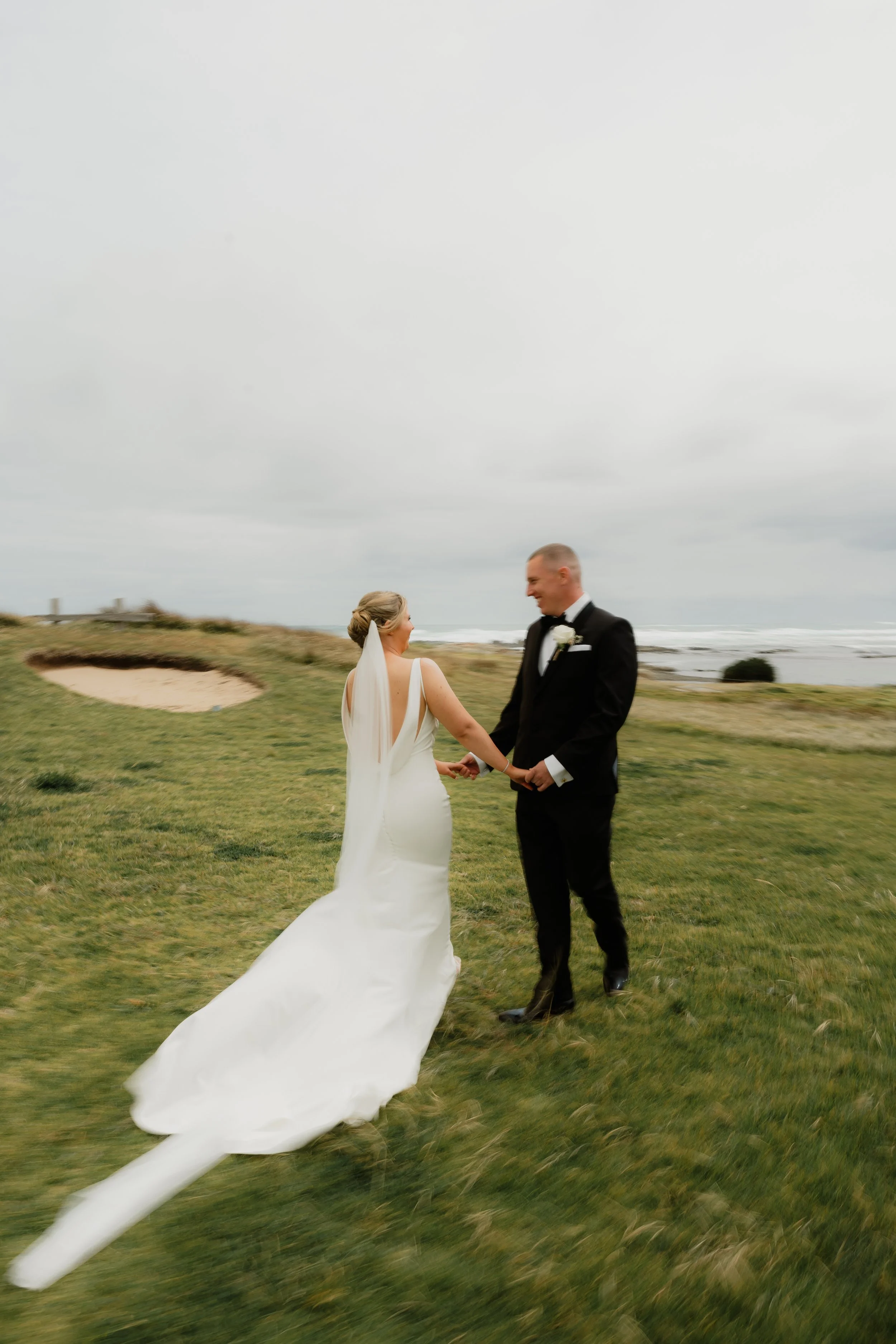 A bride and groom holding hands and smiling on a grassy outdoor area near the beach, with overcast skies in the background.