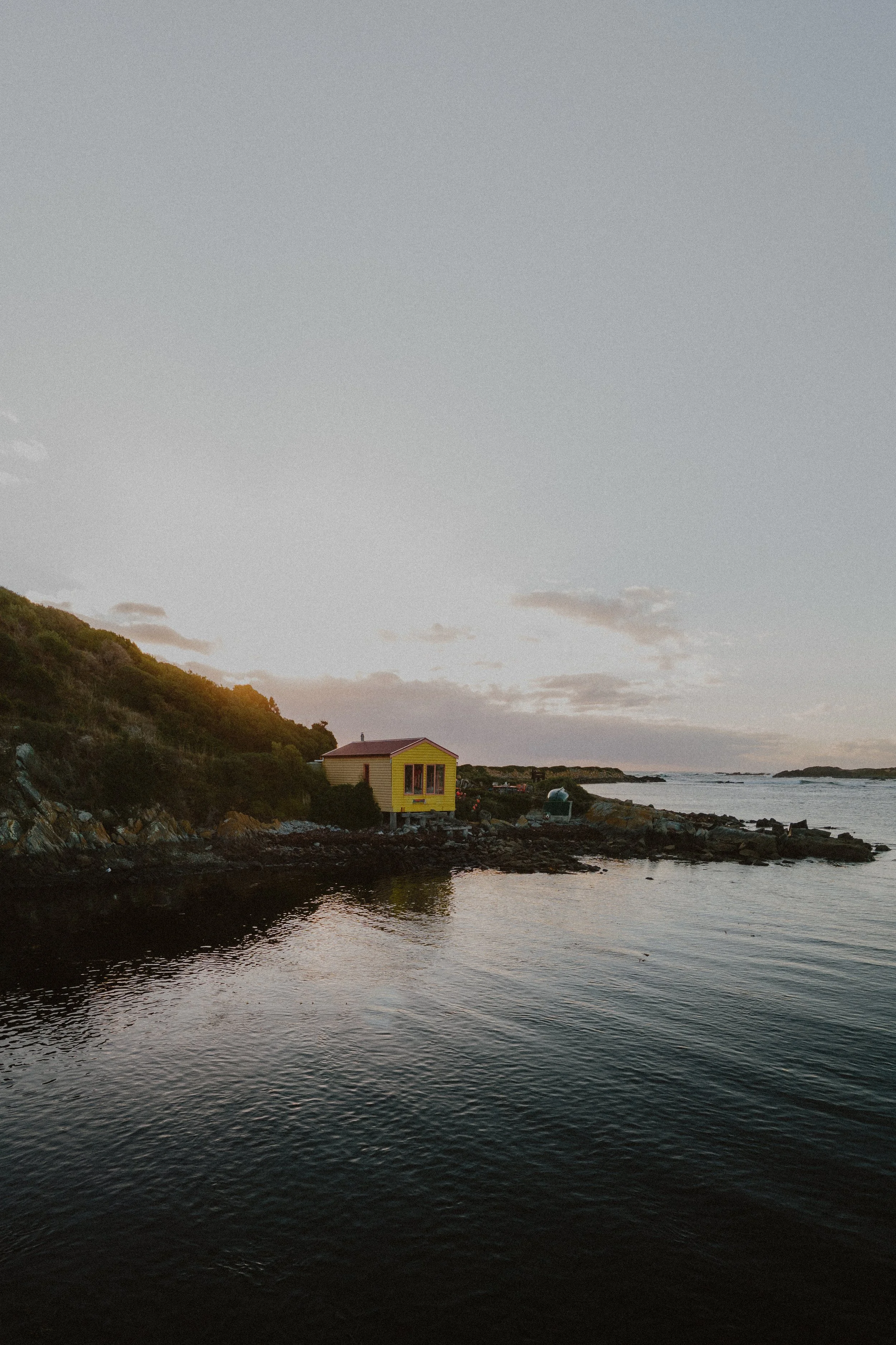 A small yellow house with a red roof sitting on rocks by the water's edge, surrounded by a hill with trees, under a partly cloudy sky during sunset.