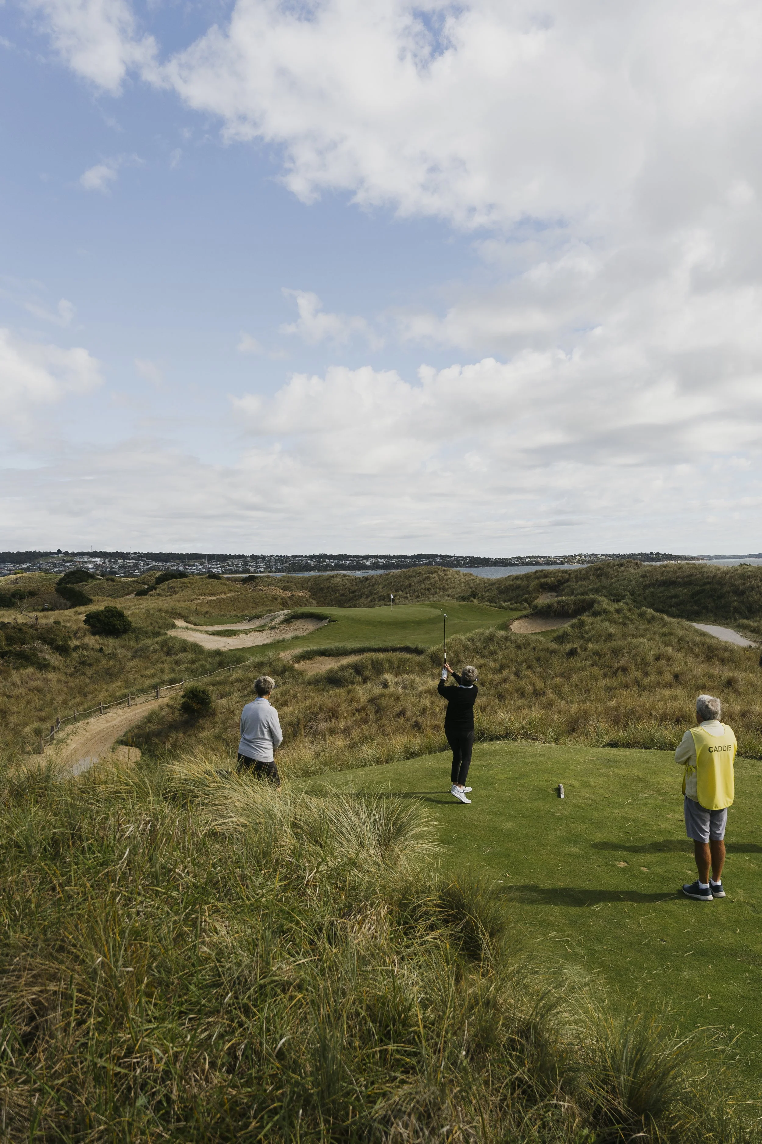 Three people on a golf course, with one swing underway and two watching, amidst grassy dunes under a partly cloudy sky.
