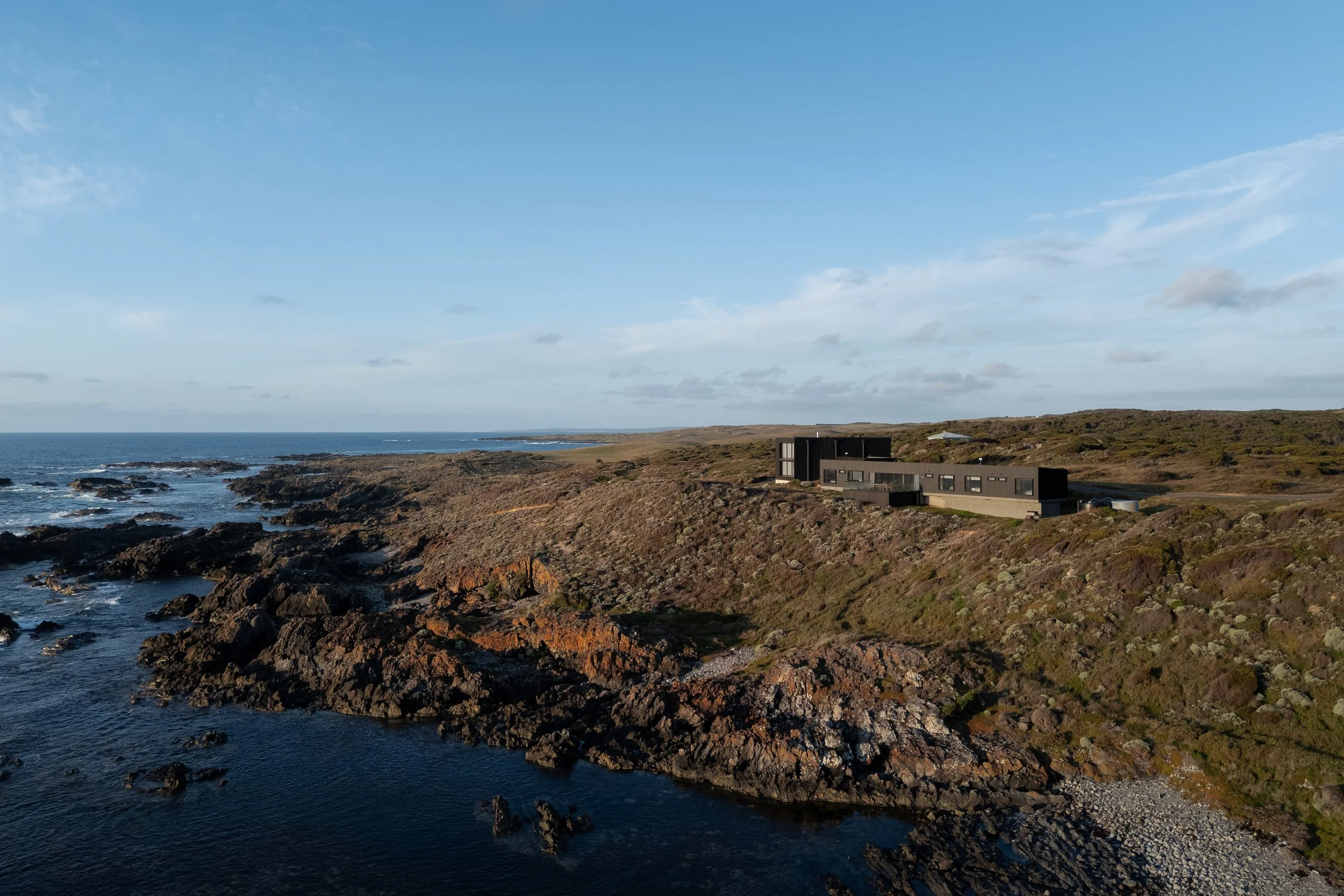 Modern black house on a rocky coastline with ocean waves and a partly cloudy sky.