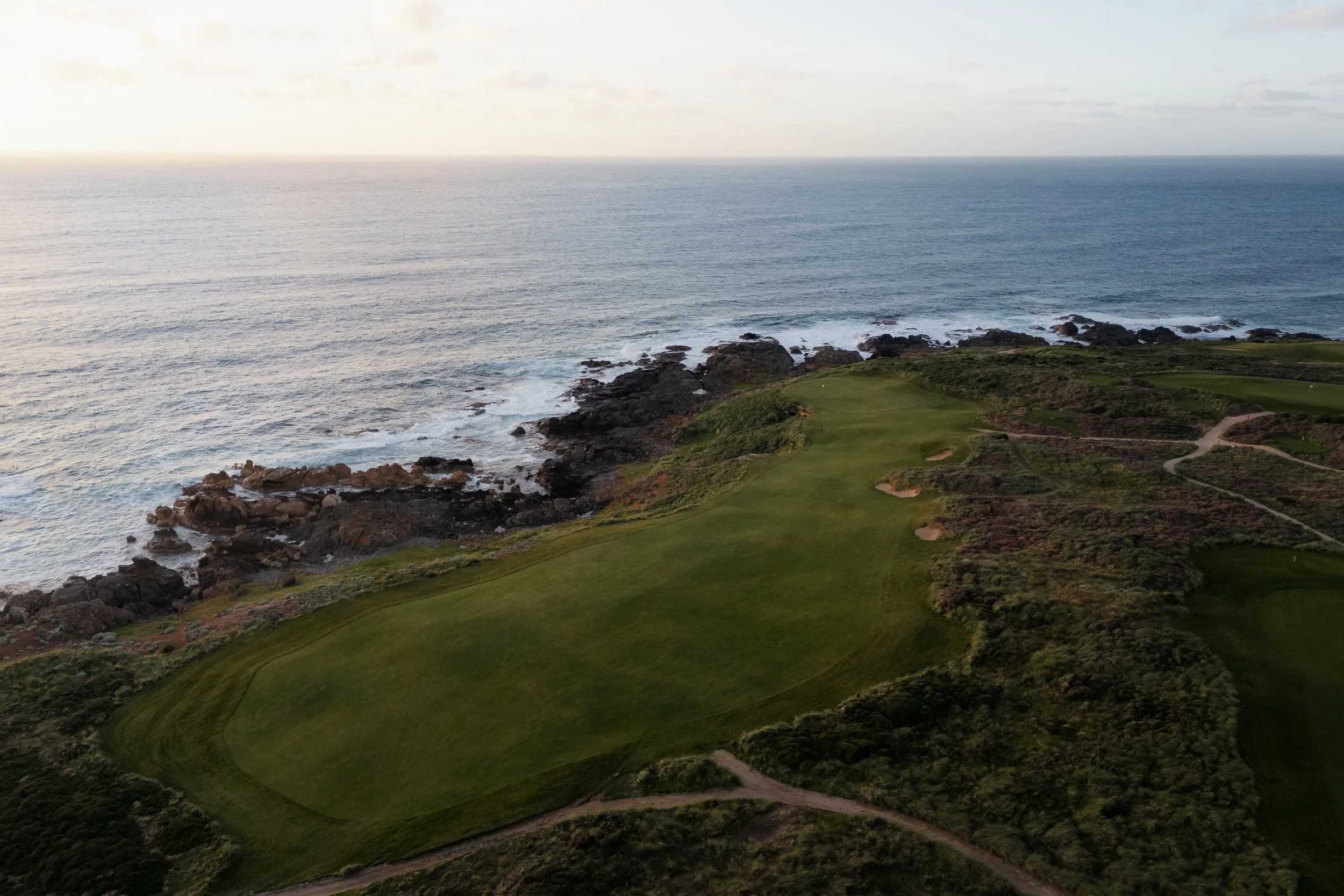 A coastal golf course with lush green fairways and rough, located on rocky cliffs, overlooking the ocean with waves hitting the rocks, under a cloudy sky.