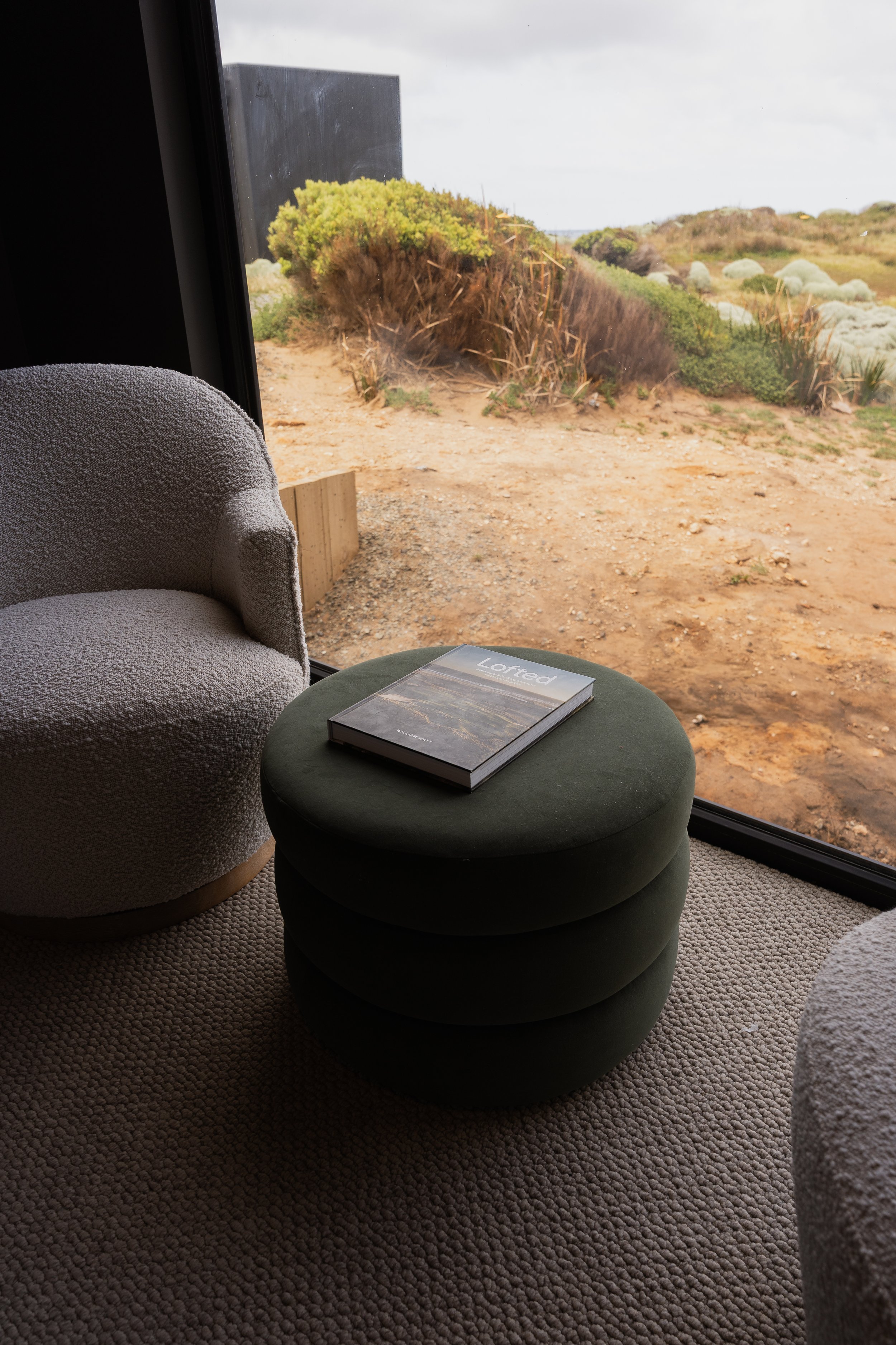 Indoor scene with a beige armchair, a green circular ottoman with a book titled "Lofed" on top, and a large window showing an outdoor landscape with dirt path, green bushes, and cloudy sky.