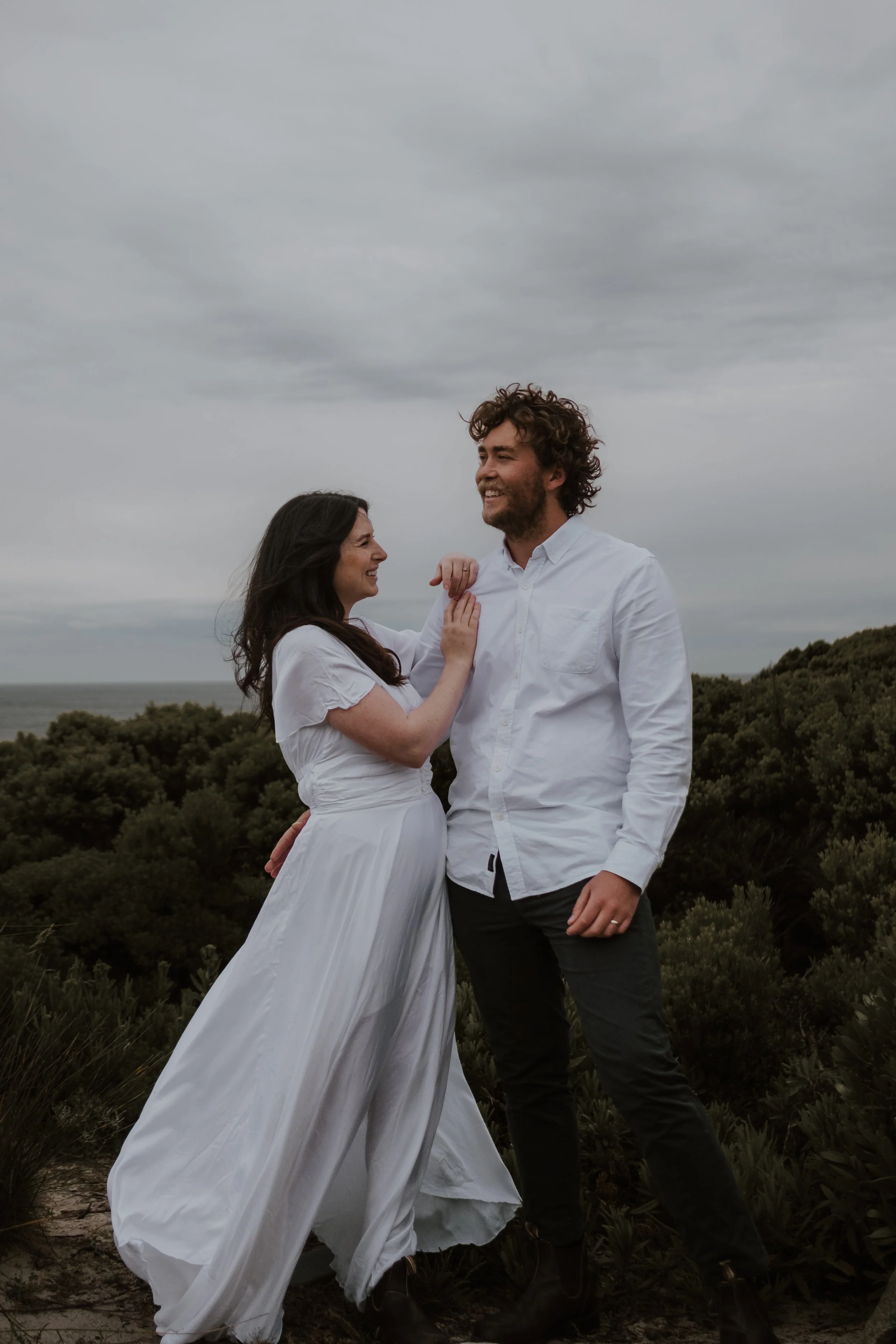 A smiling woman with dark hair wearing a white dress and a man with curly hair and beard wearing a white shirt, standing outdoors with cloudy sky and greenery in the background.