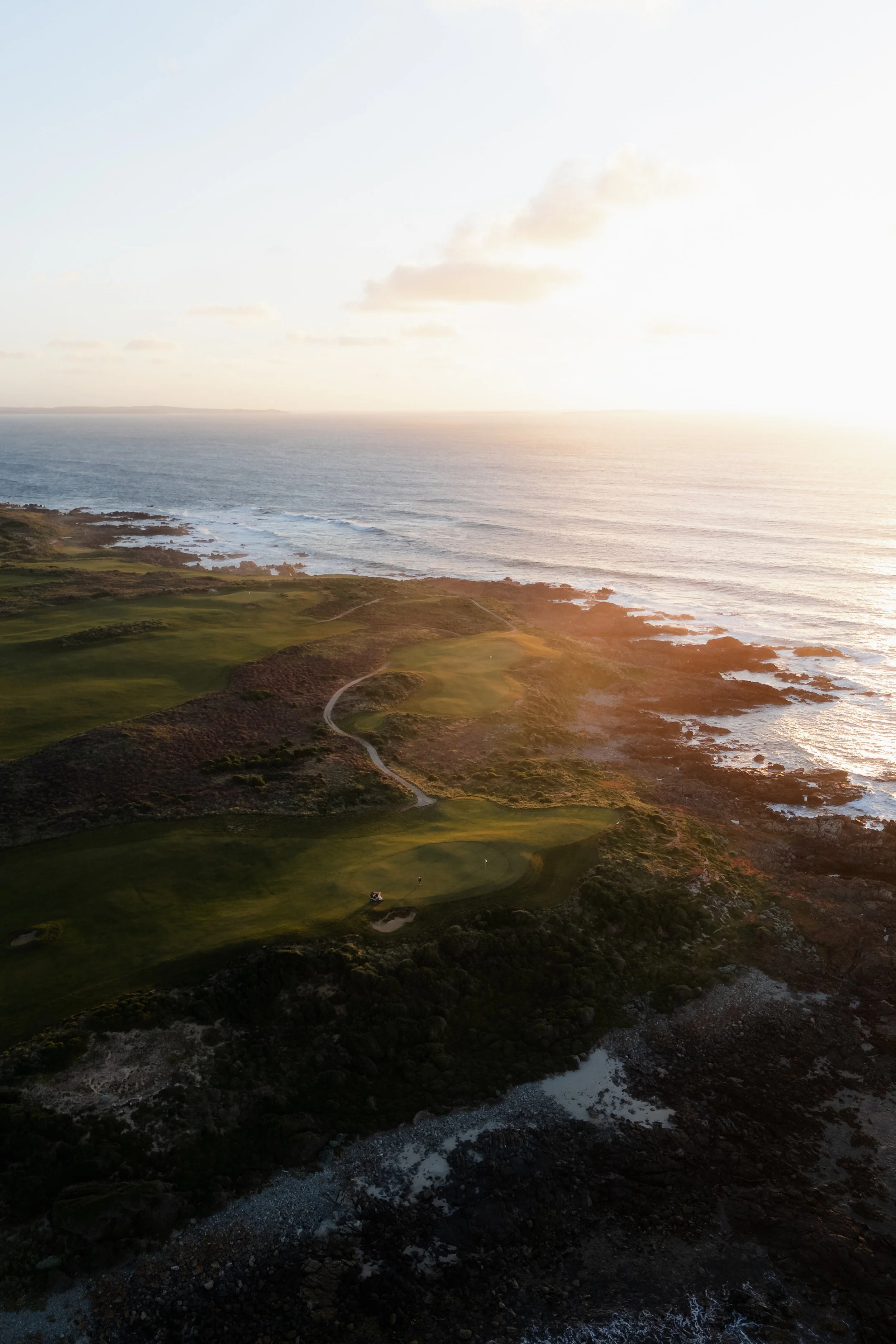 A coastal landscape with a golf course, rocky shoreline, ocean, and a sunset.