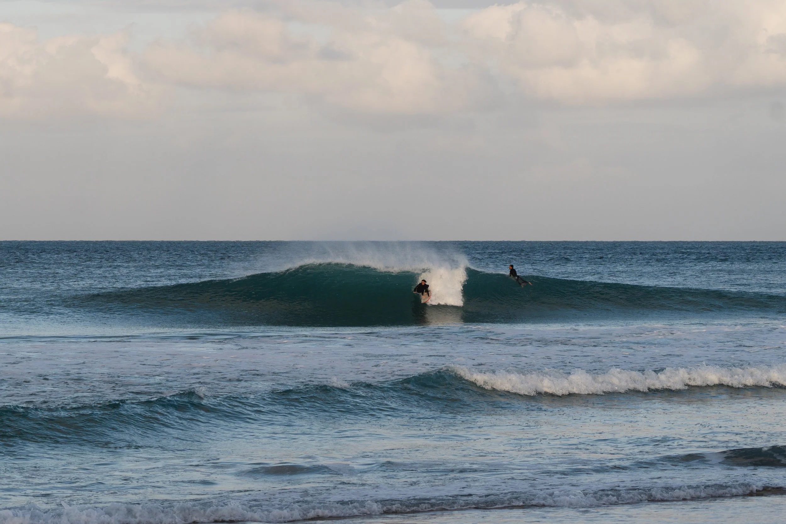 Two surfers riding a wave in the ocean, with a cloudy sky above and calm water in the foreground.