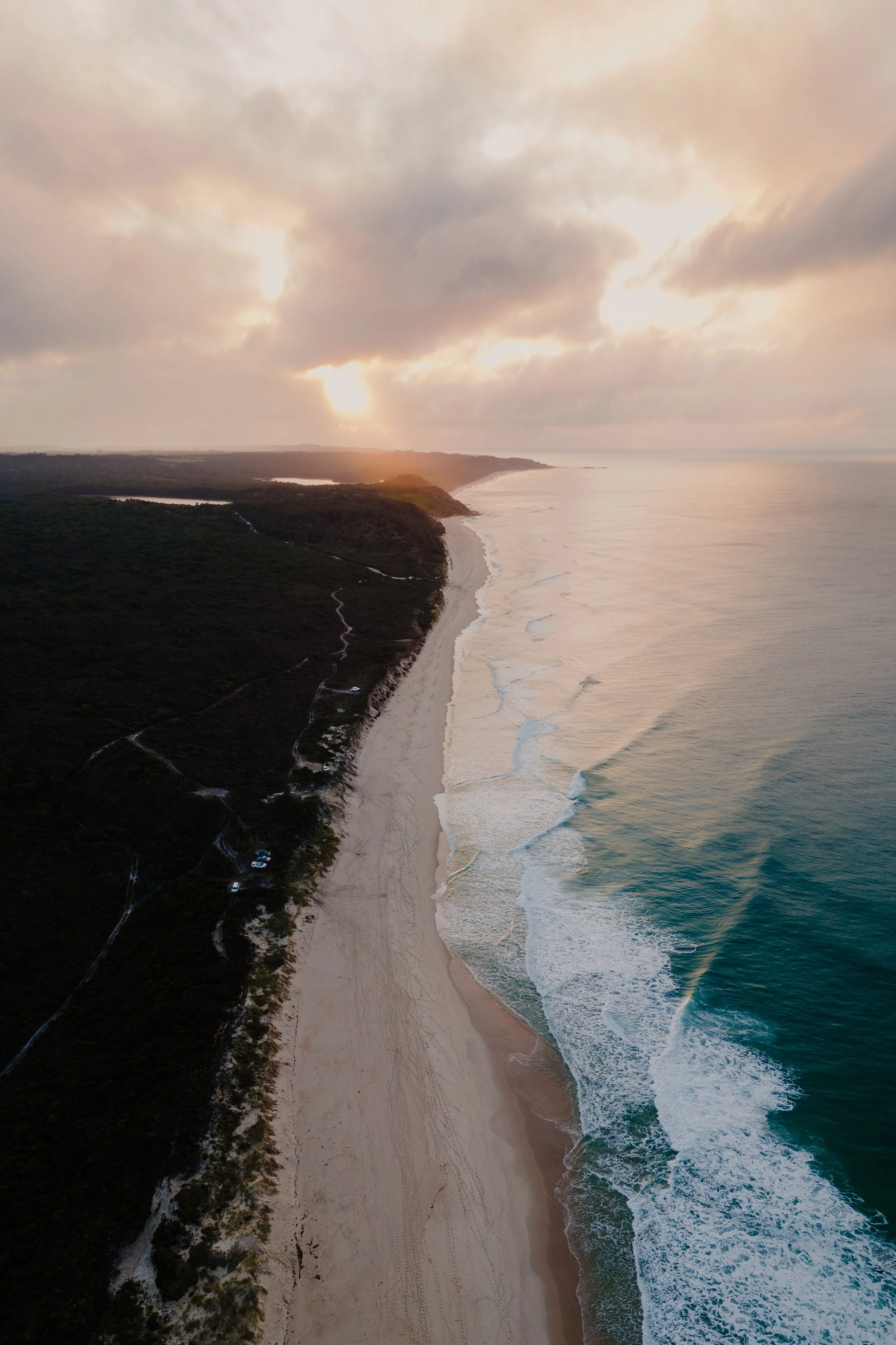 Aerial view of a sandy beach along a coastline, with green vegetation on the left and the ocean on the right, under a cloudy sky with the sun partially obscured.