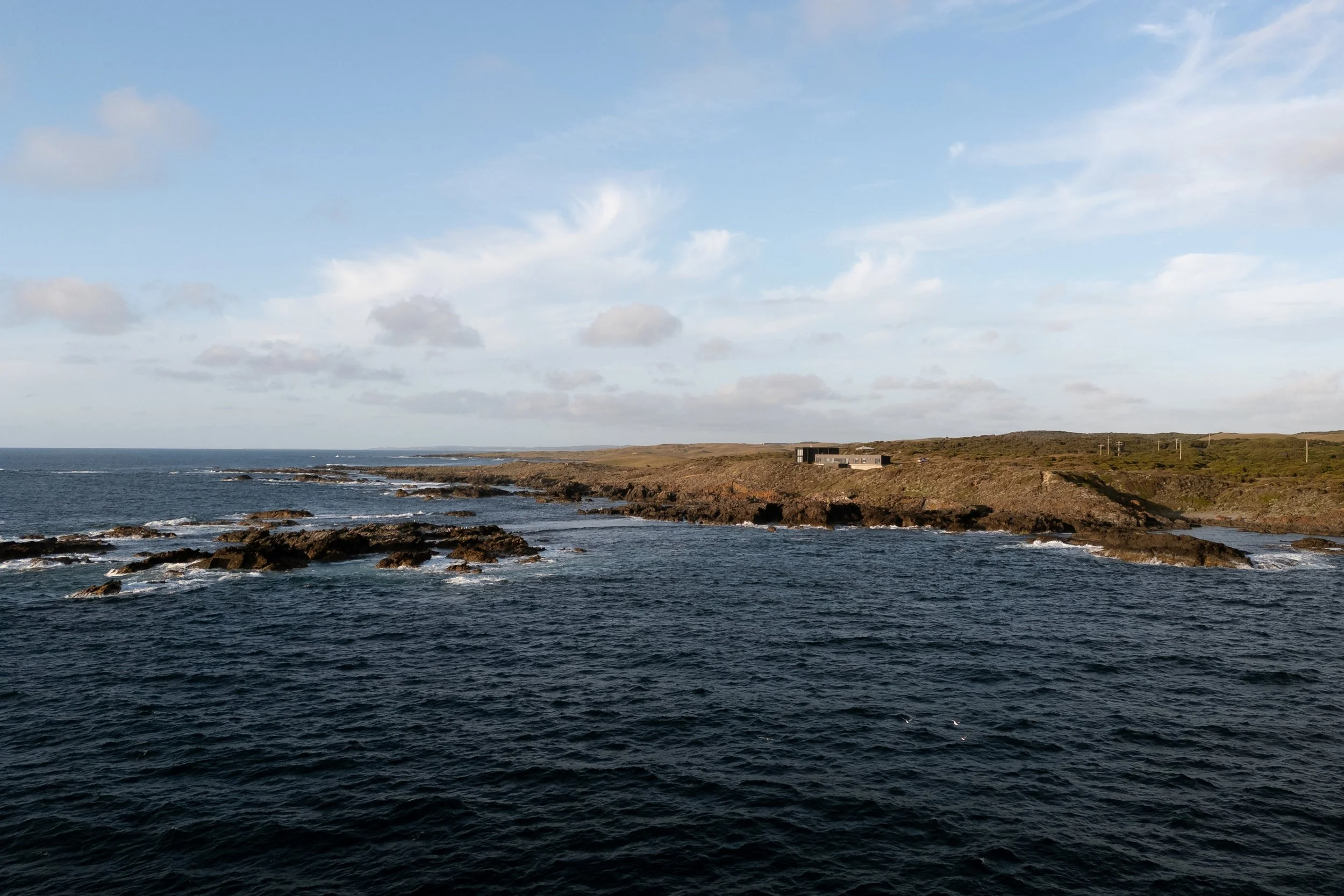 Ocean view with rocky shoreline, a grassy hillside in the distance, and a modern house on the hill under a partly cloudy sky.