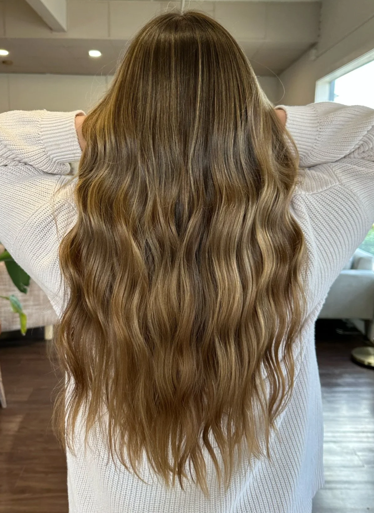 Woman with long, wavy, light brown hair seen from the back, wearing a white sweater indoors.