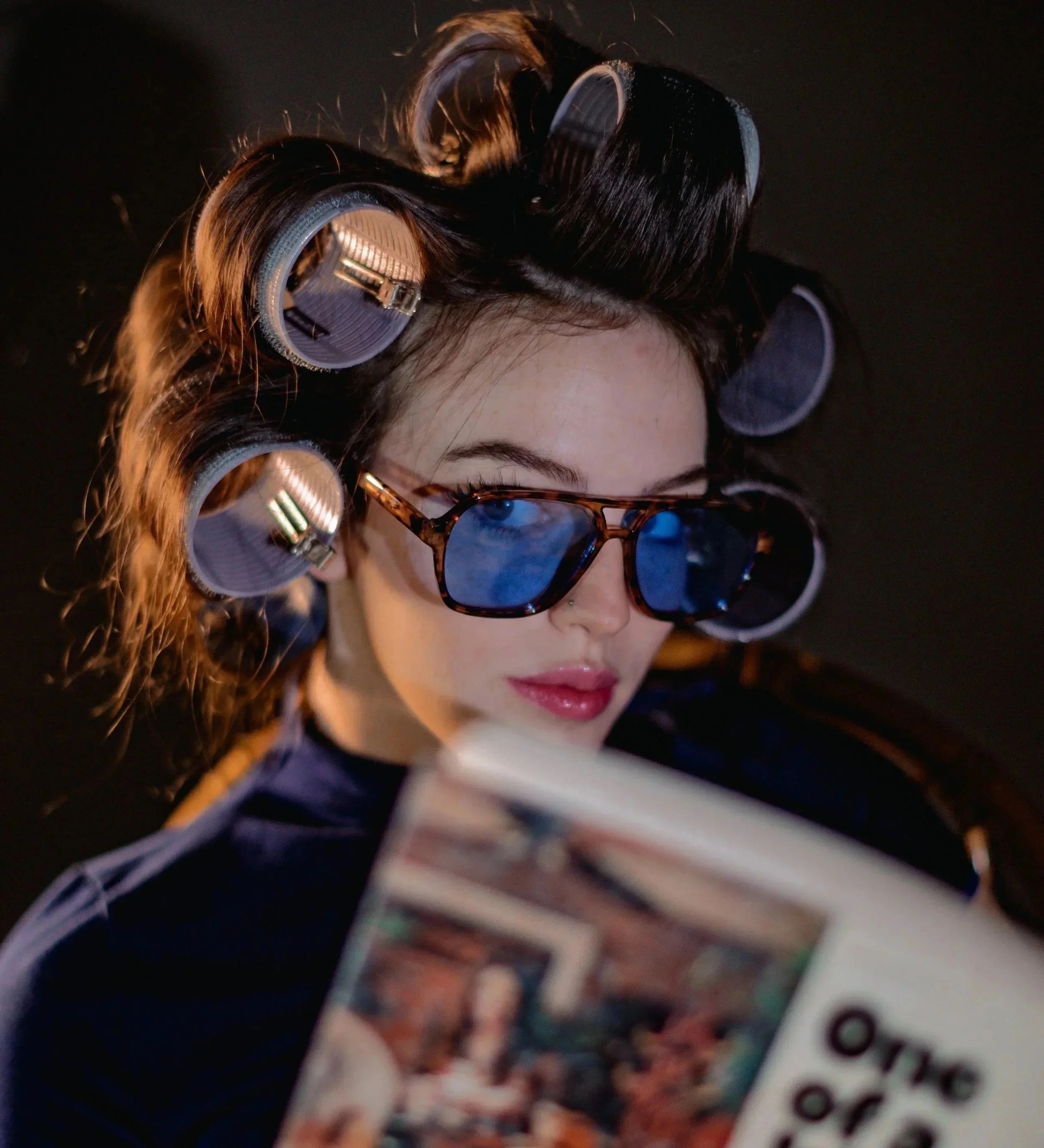 A woman with rollers in her hair and wearing sunglasses reading a magazine.