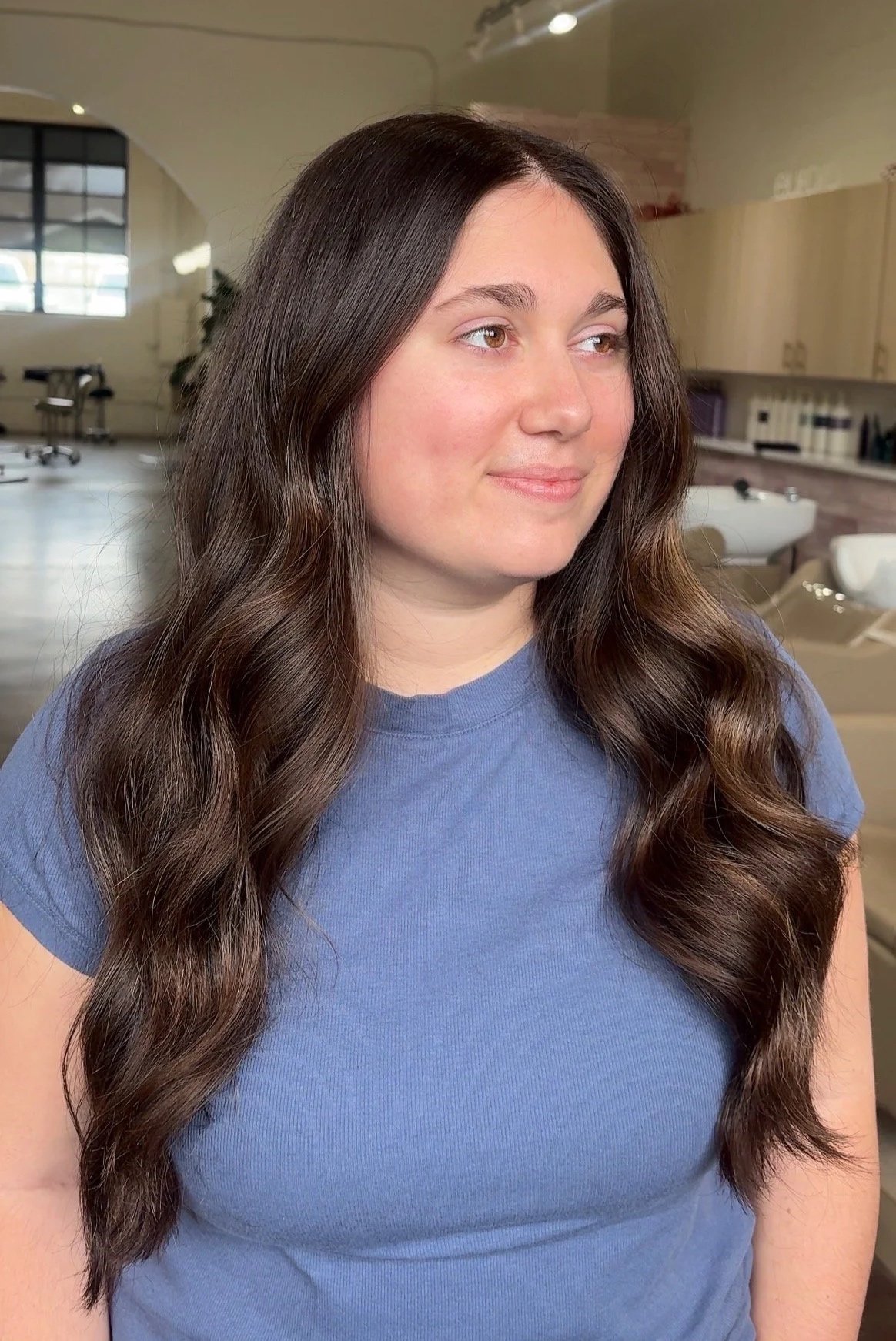 A young woman with long, wavy brown hair wearing a blue t-shirt, standing indoors in a well-lit room with large windows and beige furniture.