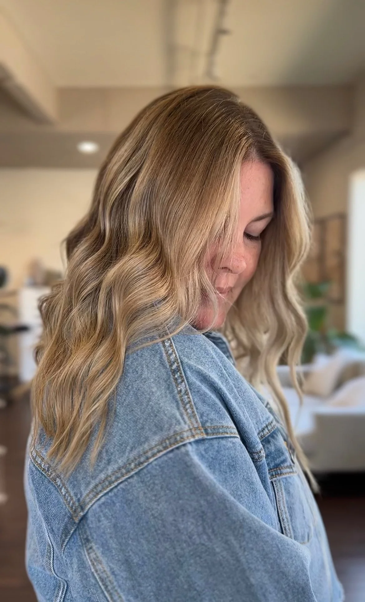 A woman with blonde wavy hair wearing a denim jacket, looking down in an indoor setting.