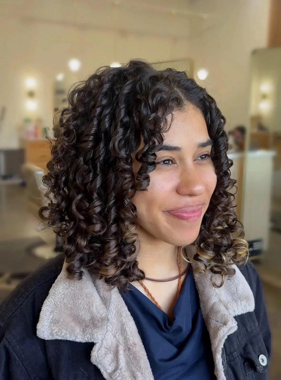 A woman with shoulder-length dark brown curly hair, light skin, and a subtle smile in a salon or similar environment.