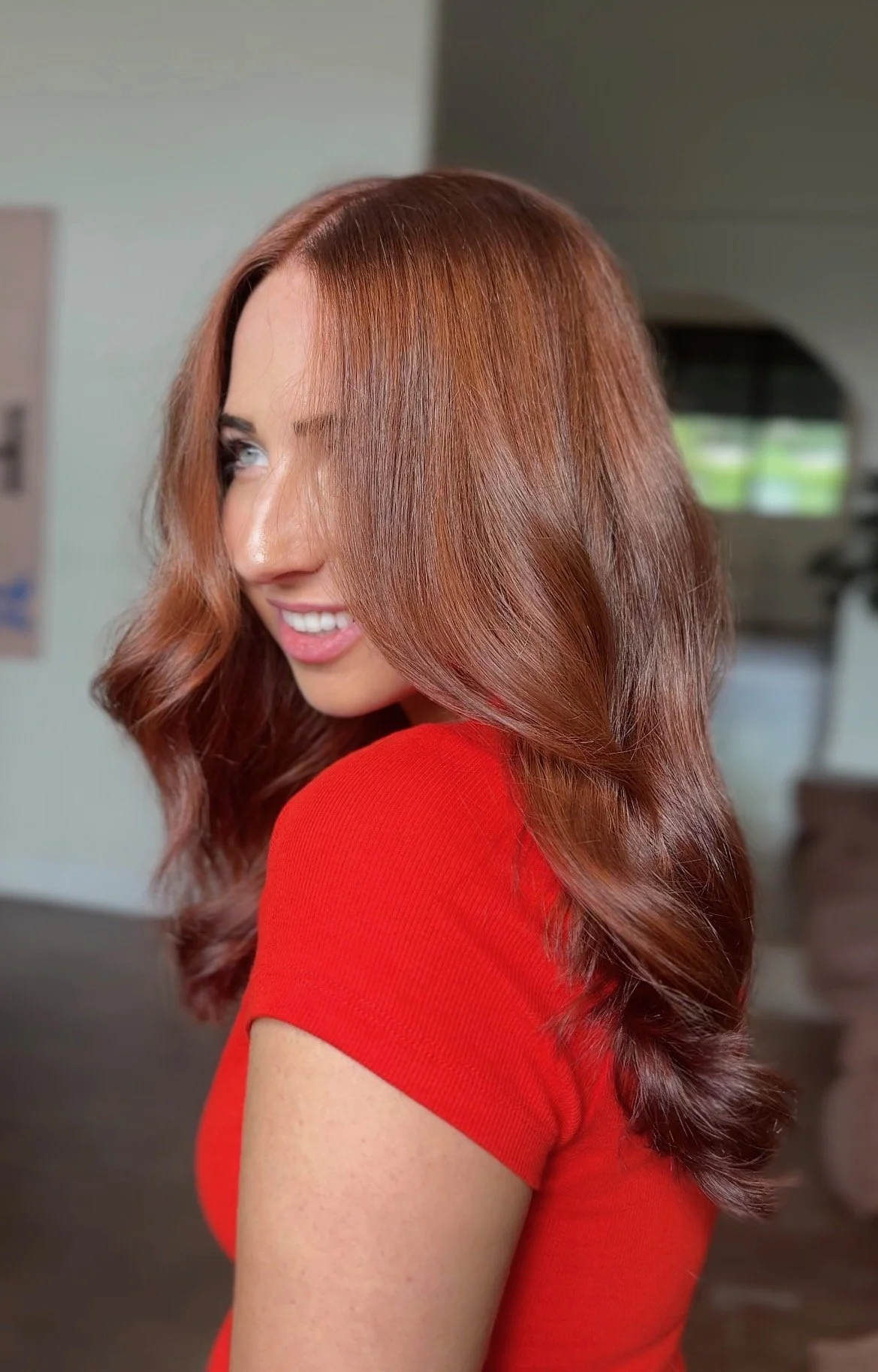 A woman with long, wavy, reddish-brown hair smiling and looking slightly away, wearing a red top inside a home.