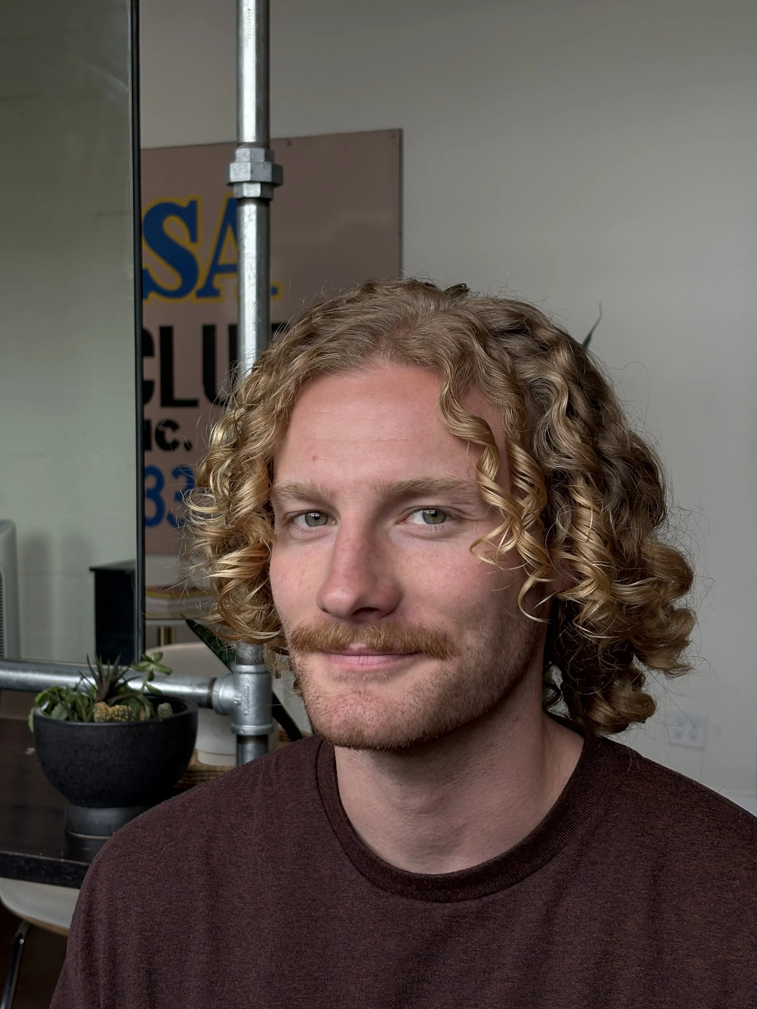 A young man with curly blonde hair, a mustache, and light skin in an indoor setting, smiling slightly at the camera.