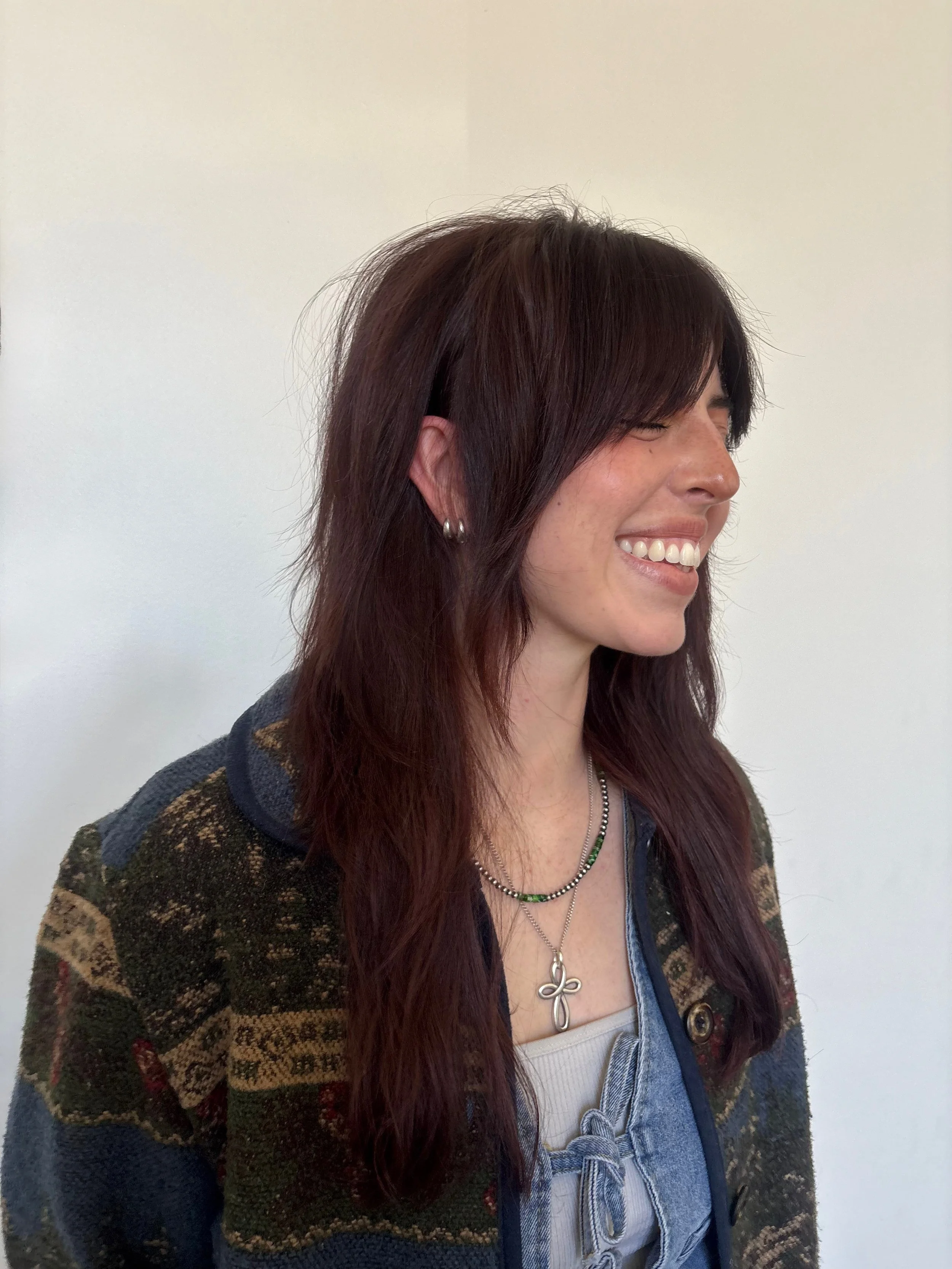 A woman with long reddish-brown hair, styled with bangs, smiling and showing white teeth, wearing a patterned sweater and layered necklaces, standing against a plain light-colored wall.
