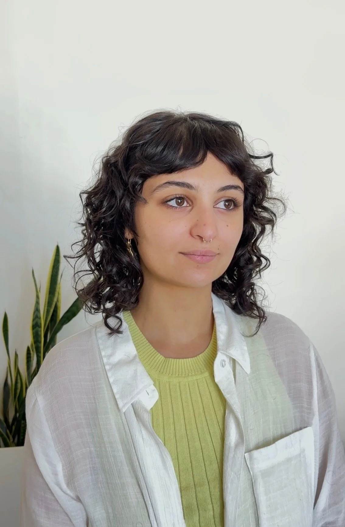 A young woman with dark, curly hair, light skin, and brown eyes, wearing a white button-up shirt over a light green ribbed top, sitting near a plant with long, green leaves against a plain light-colored wall.