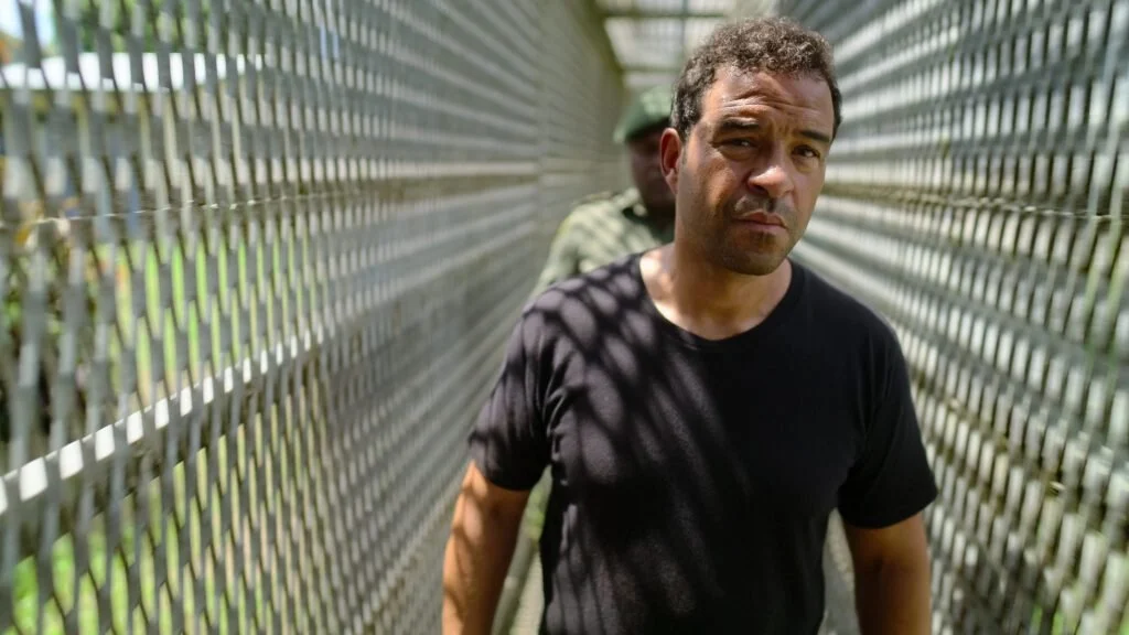 A man walking along a narrow, metal fence walkway in a prison, with a guard behind him.