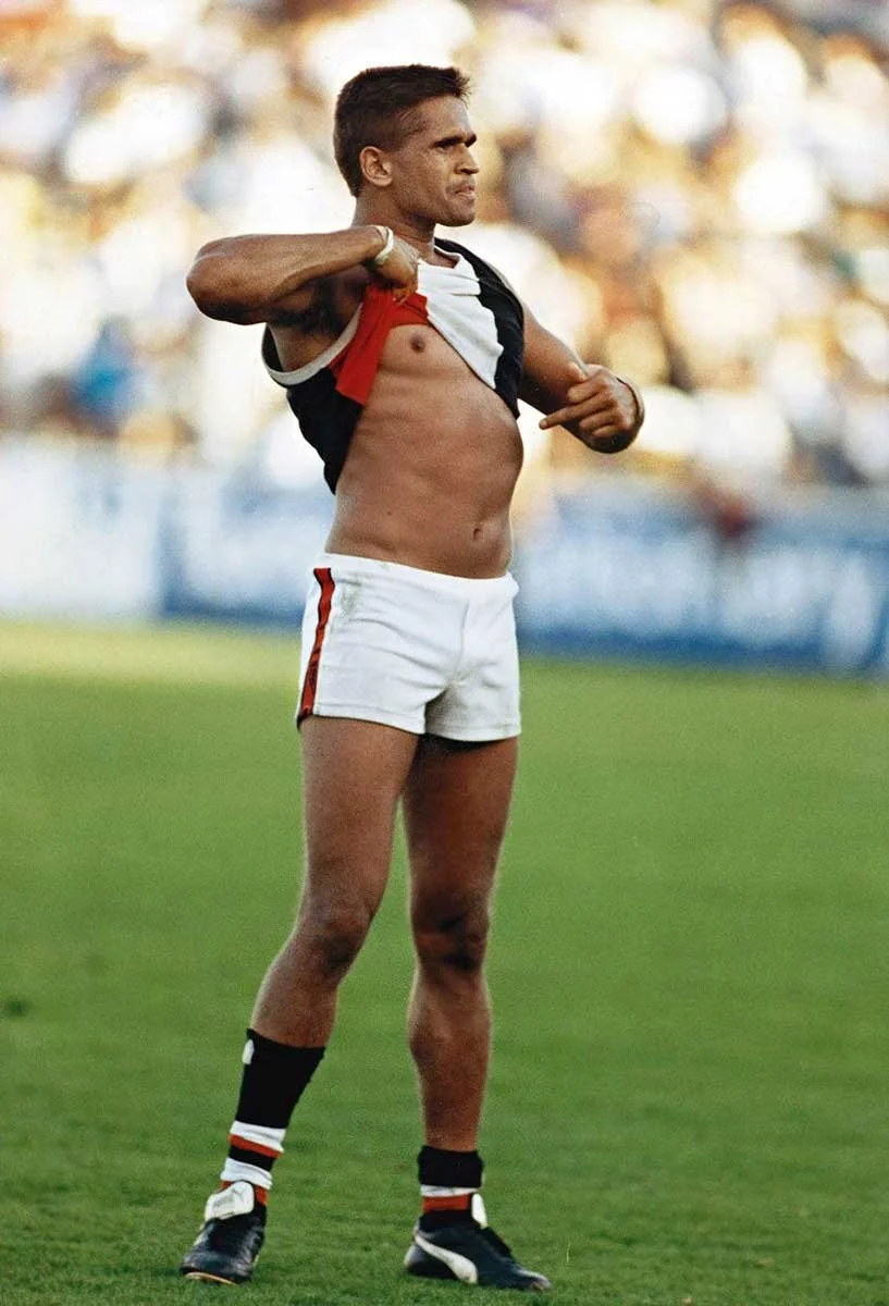 Noongar man, Nicky Winmar is on a football field, lifting his jersey to expose his Aboriginal skin with a focused, determined expression he points to his chest.