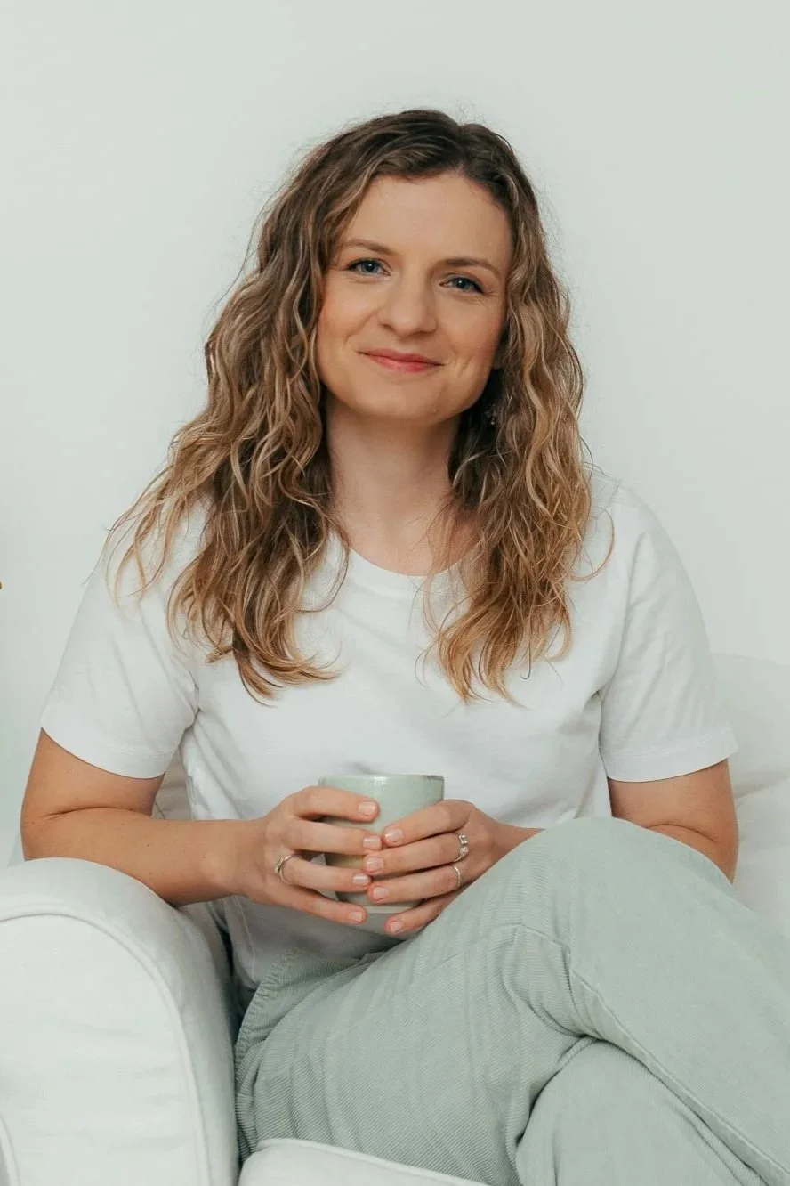 A woman with curly hair sitting on a white couch, holding a mug, wearing a white t-shirt and light-colored pants, smiling at the camera.