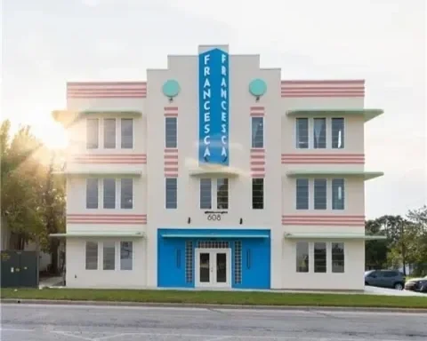 Art Deco style building with a blue vertical sign reading 'FRANCESCA' and a central entrance, surrounded by a lawn and trees.
