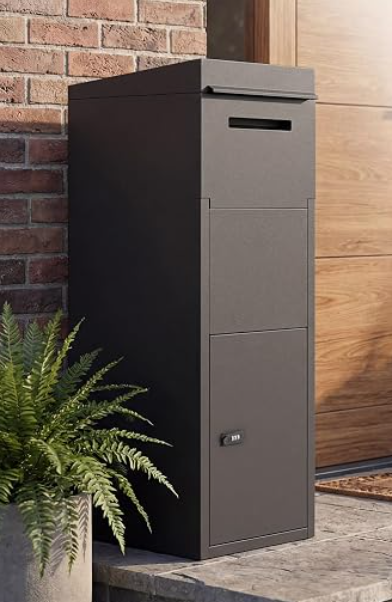 A tall, black mailbox with a slot for mail and a lockable compartment, placed outside on a concrete surface next to a potted fern plant.