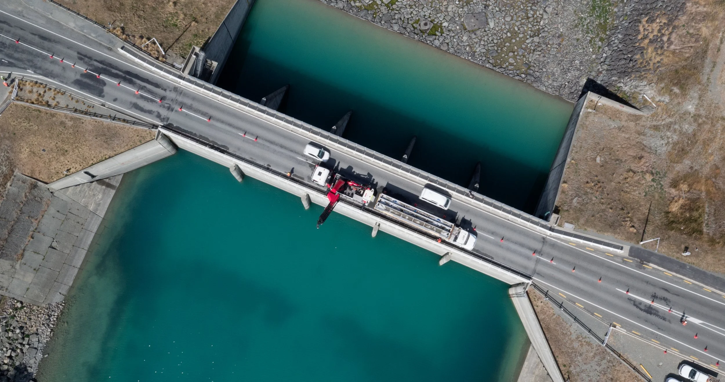 Aerial view of a dam with water on both sides and a bridge with vehicles, including trucks and cars, crossing over it.